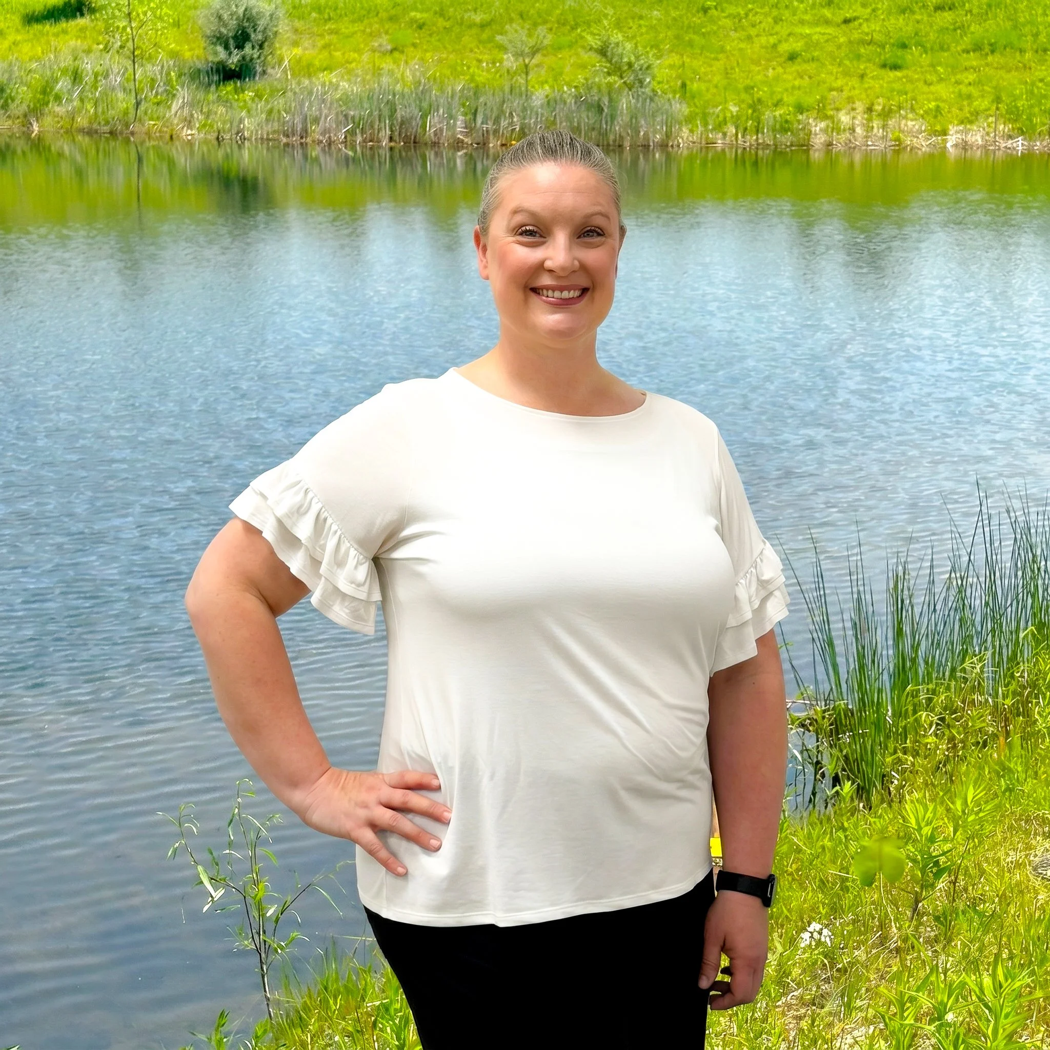 A woman smiling by a lake with green grass and plants around the water.