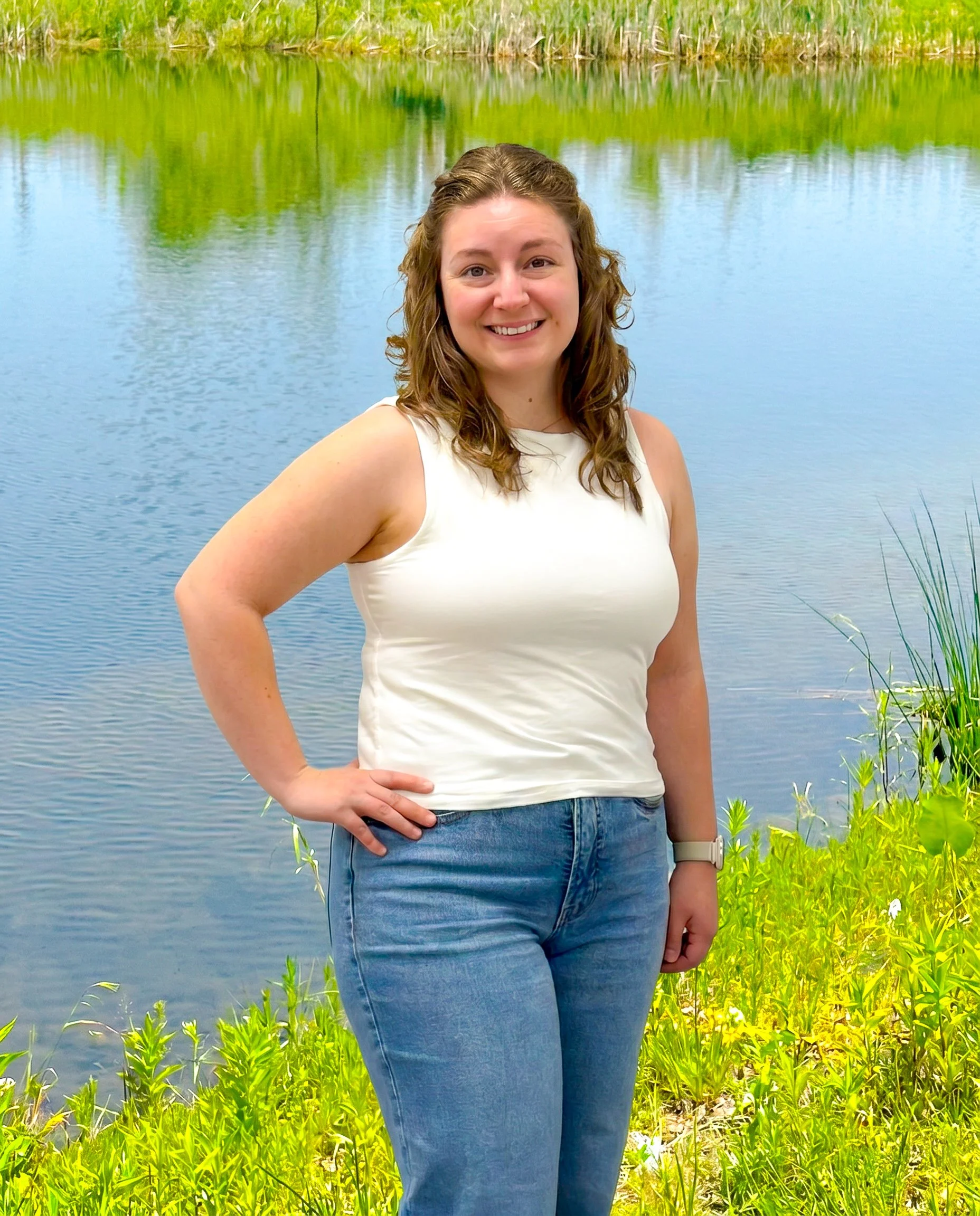 Smiling woman with light brown hair wearing a white sleeveless top and blue jeans stands by a lake with green plants and trees reflected in the water.