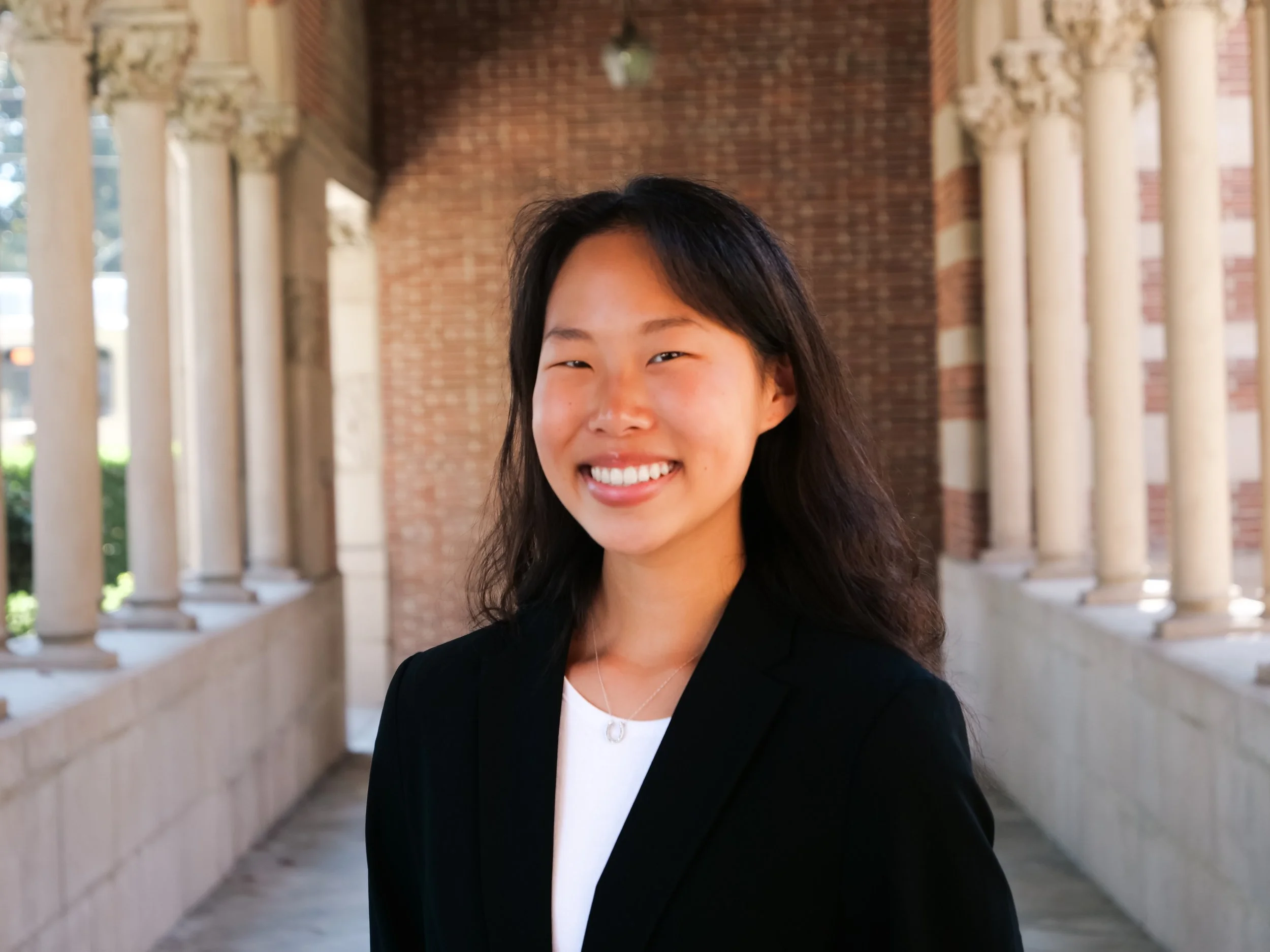 A young woman smiling outdoors in a professional setting with columns and a brick building in the background.