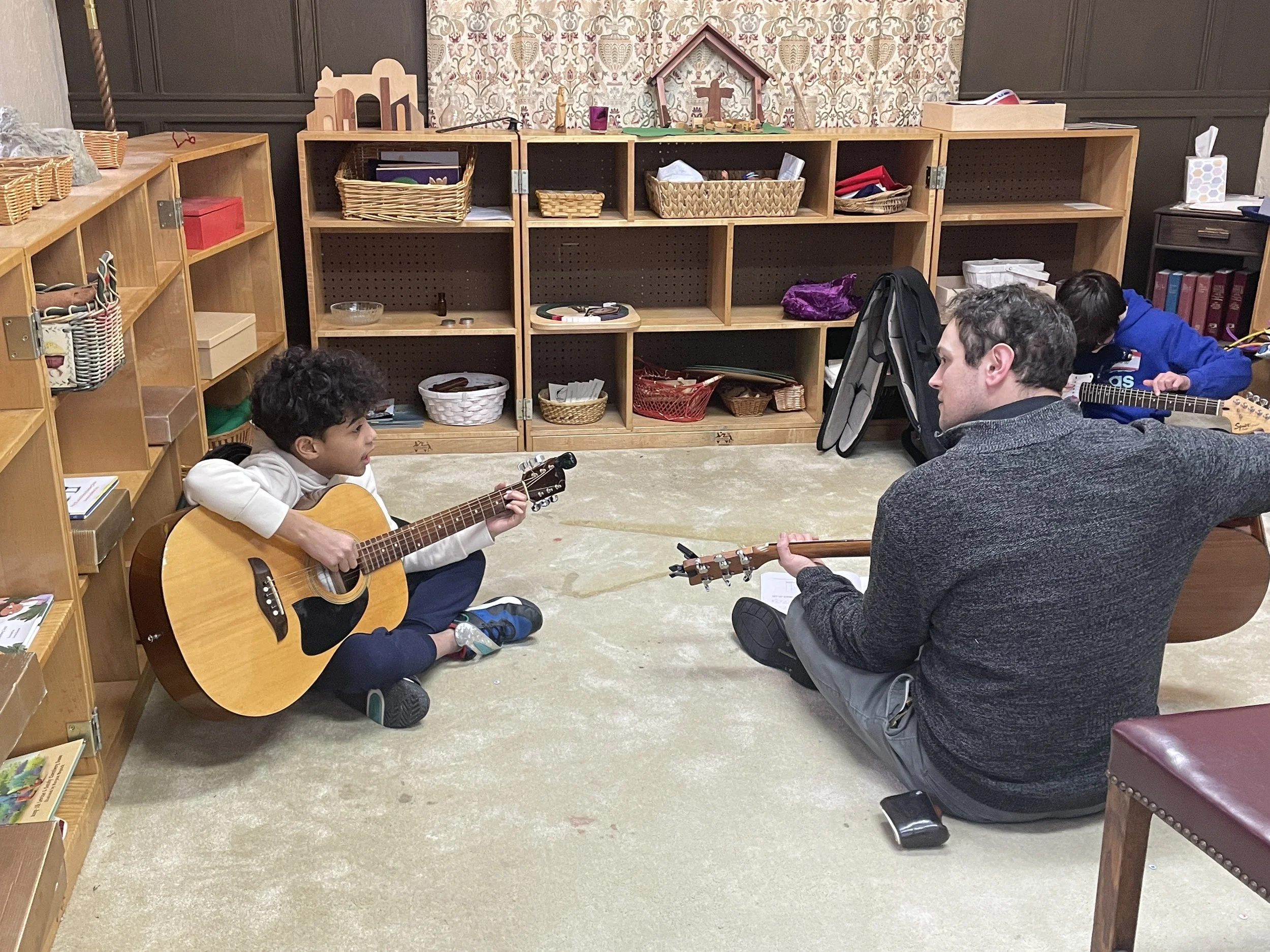 A music lesson taking place with a young boy and an adult man sitting on the floor, each holding guitars, in a room with shelves filled with books and baskets.