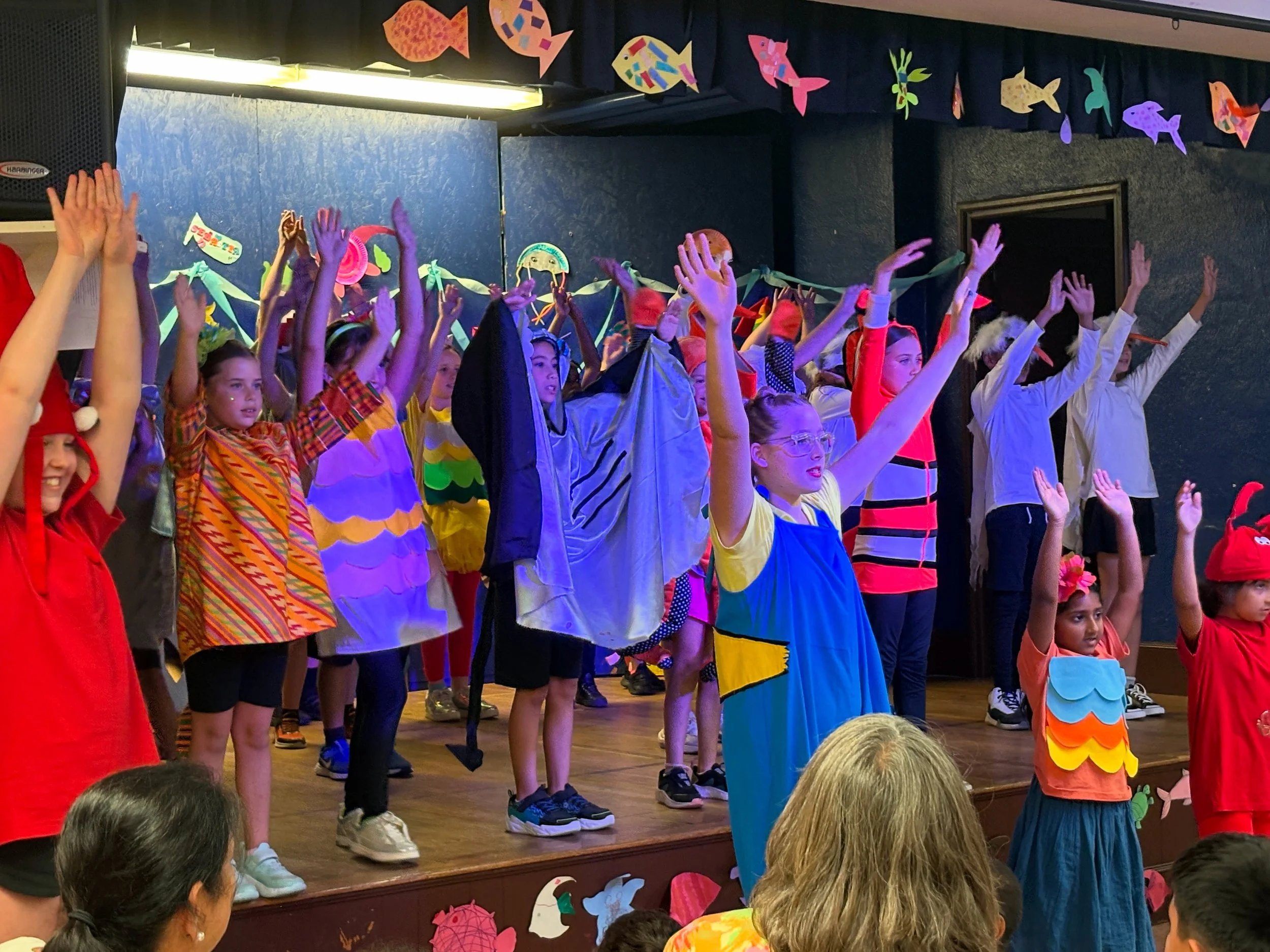 Children performing on a decorated stage, dressed in colorful costumes, with ocean-themed decorations and fish hanging from the ceiling.