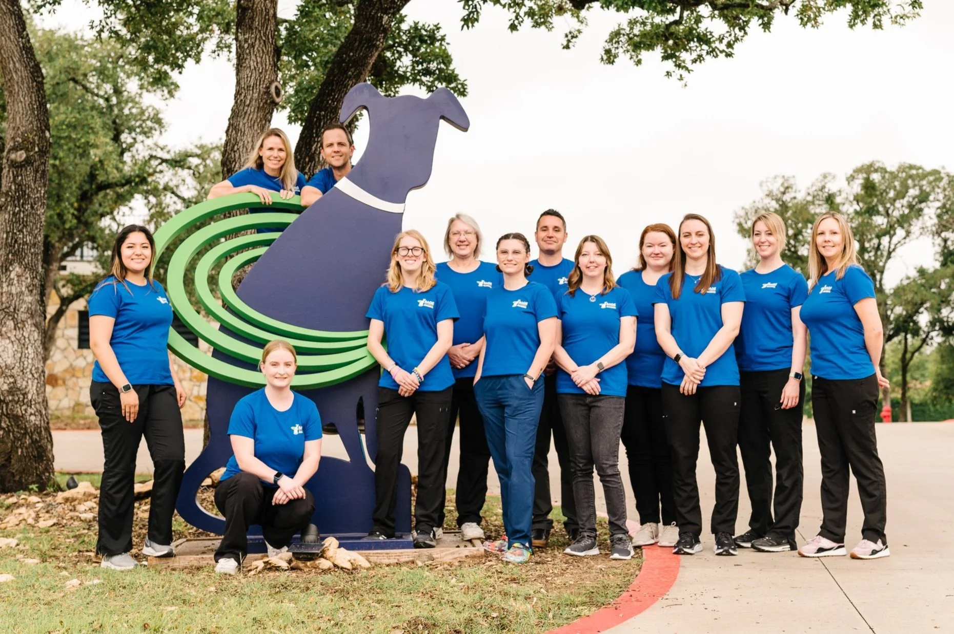 Group photo of the Sage Veterinary Imaging team in blue shirts standing beside a dog-shaped clinic sign outdoors.
