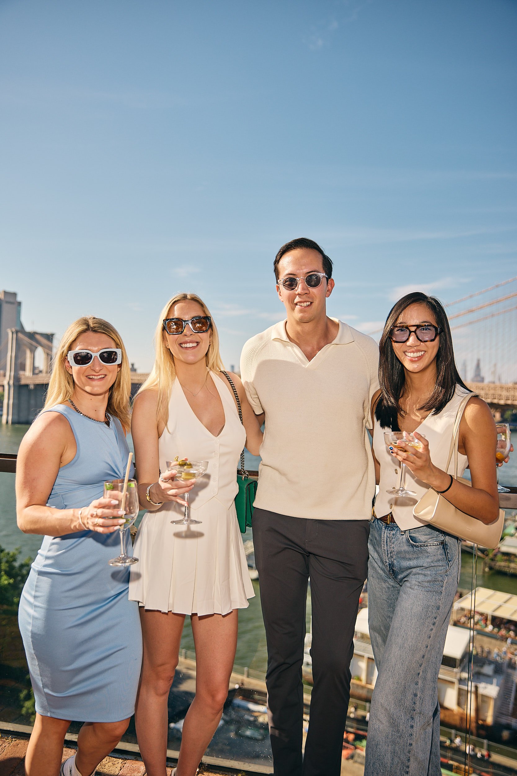 Four friends enjoying drinks on a rooftop with a bridge and city skyline in the background on a sunny day.