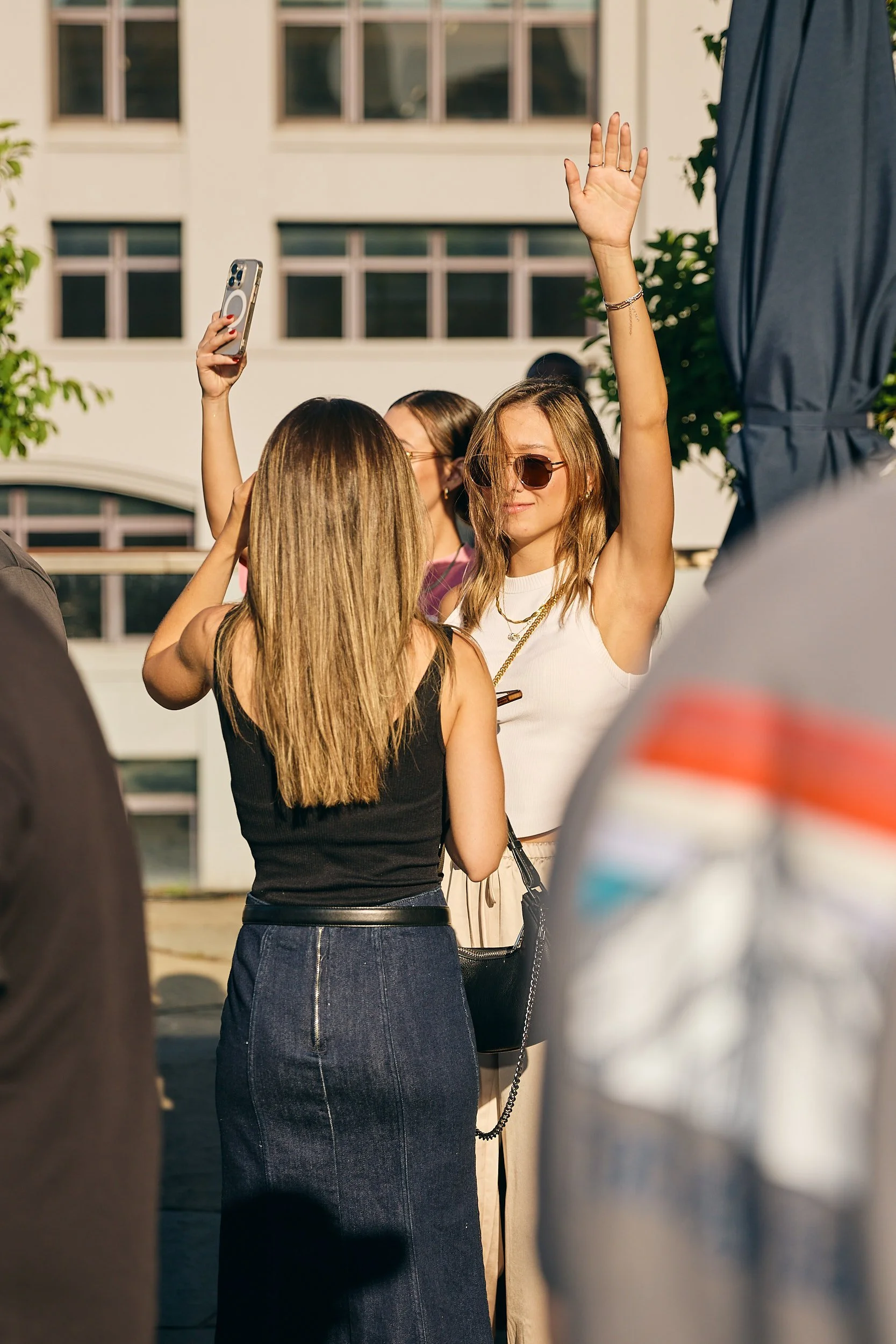 A group of young women at an outdoor social gathering, one woman with long light brown hair is taking a selfie with two other women, one with long blonde hair and another with light brown hair and sunglasses, in front of a building during sunny weath
