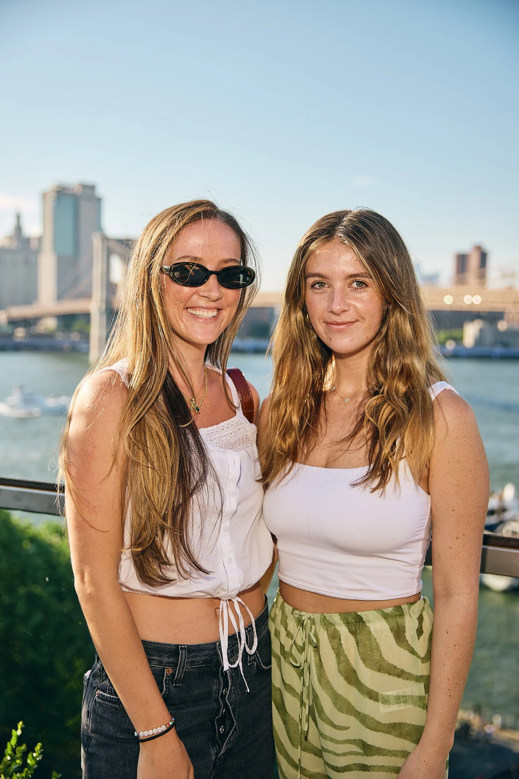 Two women are standing outdoors near a river, with a city skyline and a bridge in the background on a sunny day.