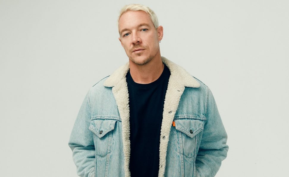 A young man with platinum blonde hair, wearing a light blue denim jacket with a shearling lining, standing against a plain background.