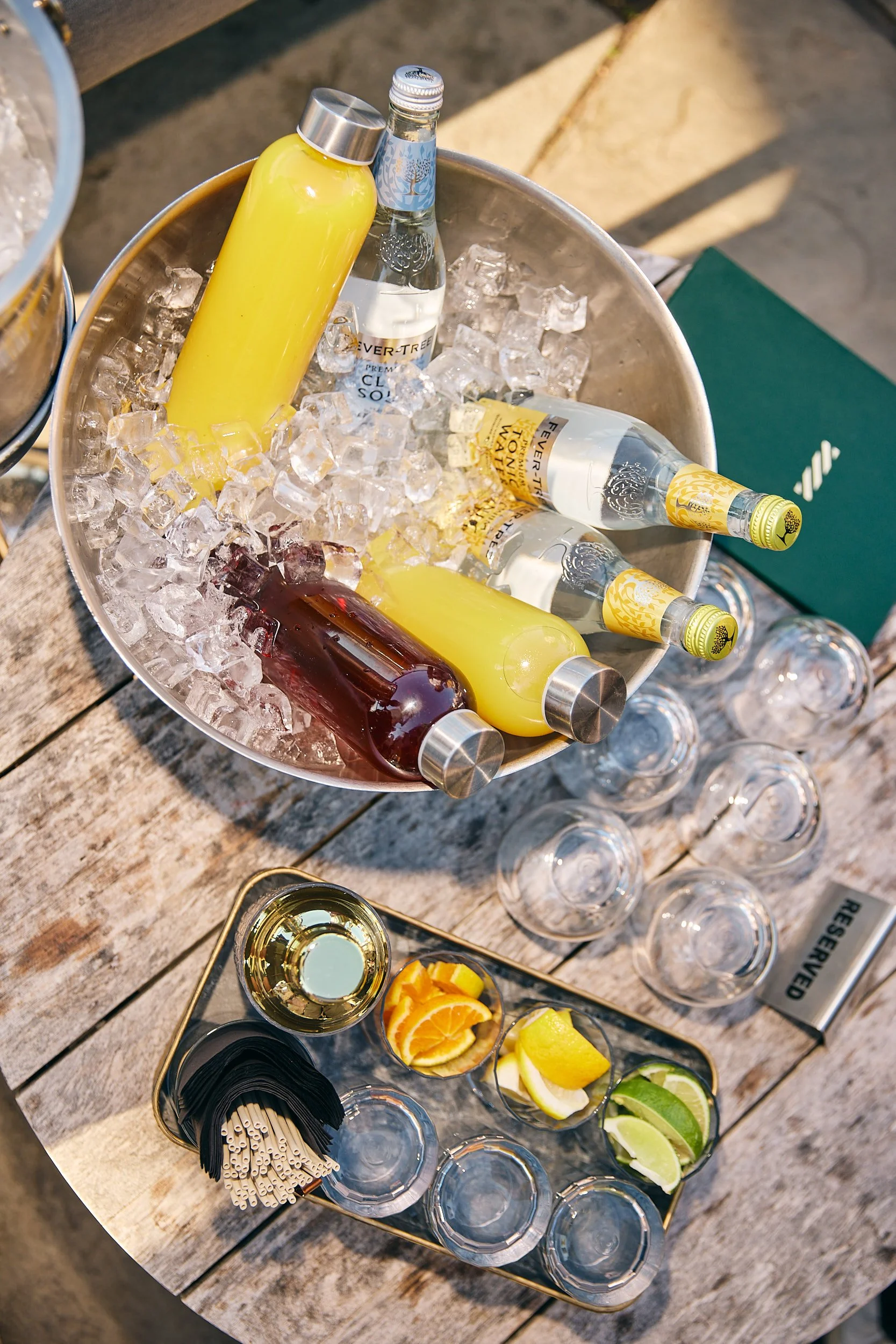 Beverage station with bottles of flavored water, glasses, and lemon, lime, and orange slices on a wooden table outdoors.