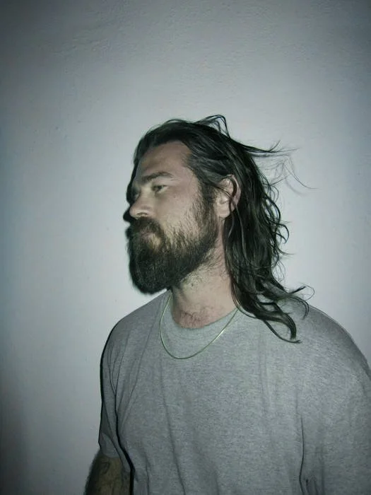 A man with long, dark hair and a beard is standing against a plain wall, wearing a gray t-shirt and a thin gold chain necklace.