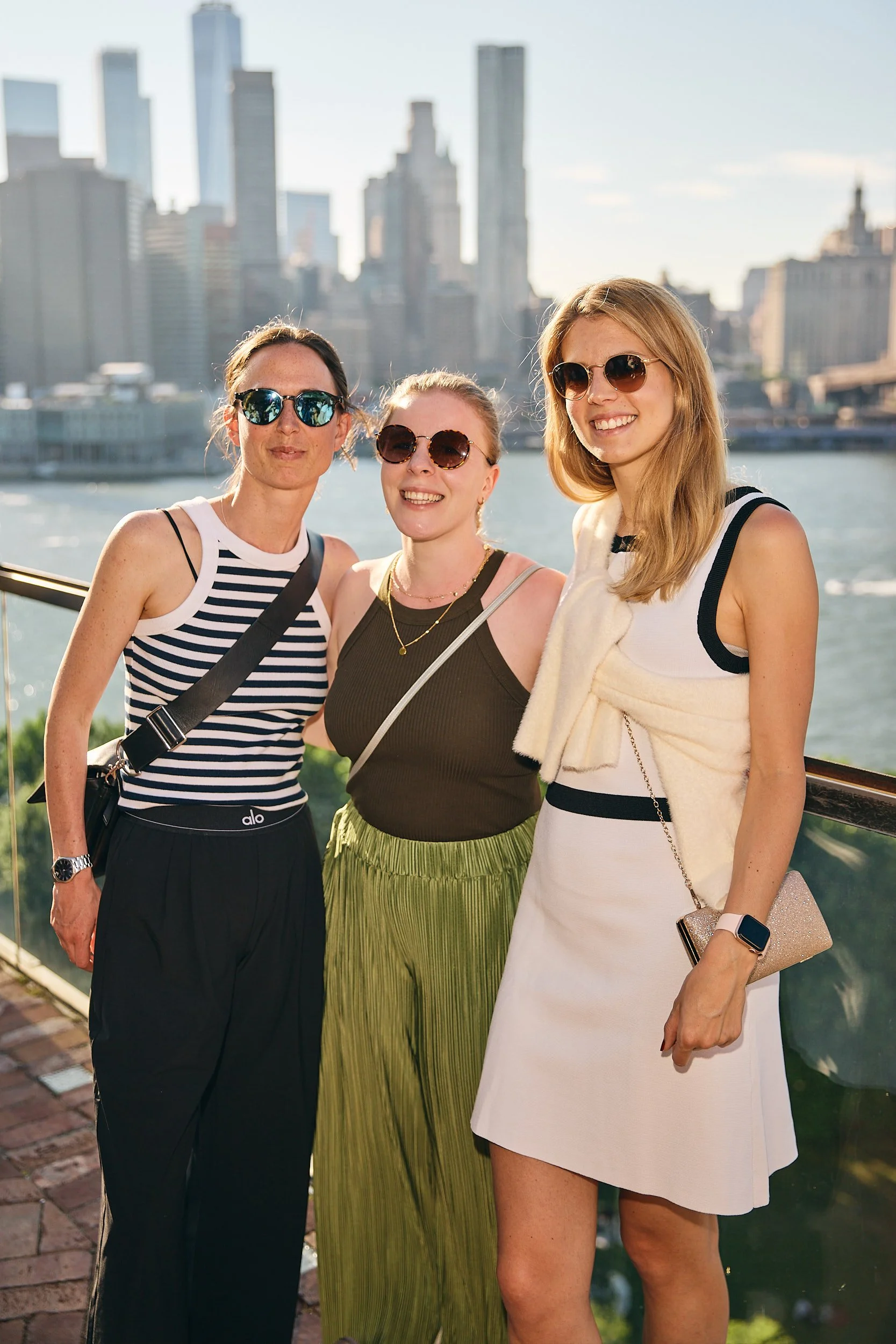 Three women smiling outdoors near a river with a city skyline in the background.