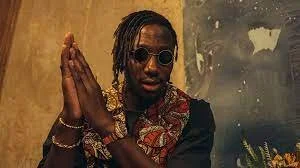 Man wearing sunglasses and a patterned shirt, holding hands together in a prayer-like pose, standing against a textured wall.