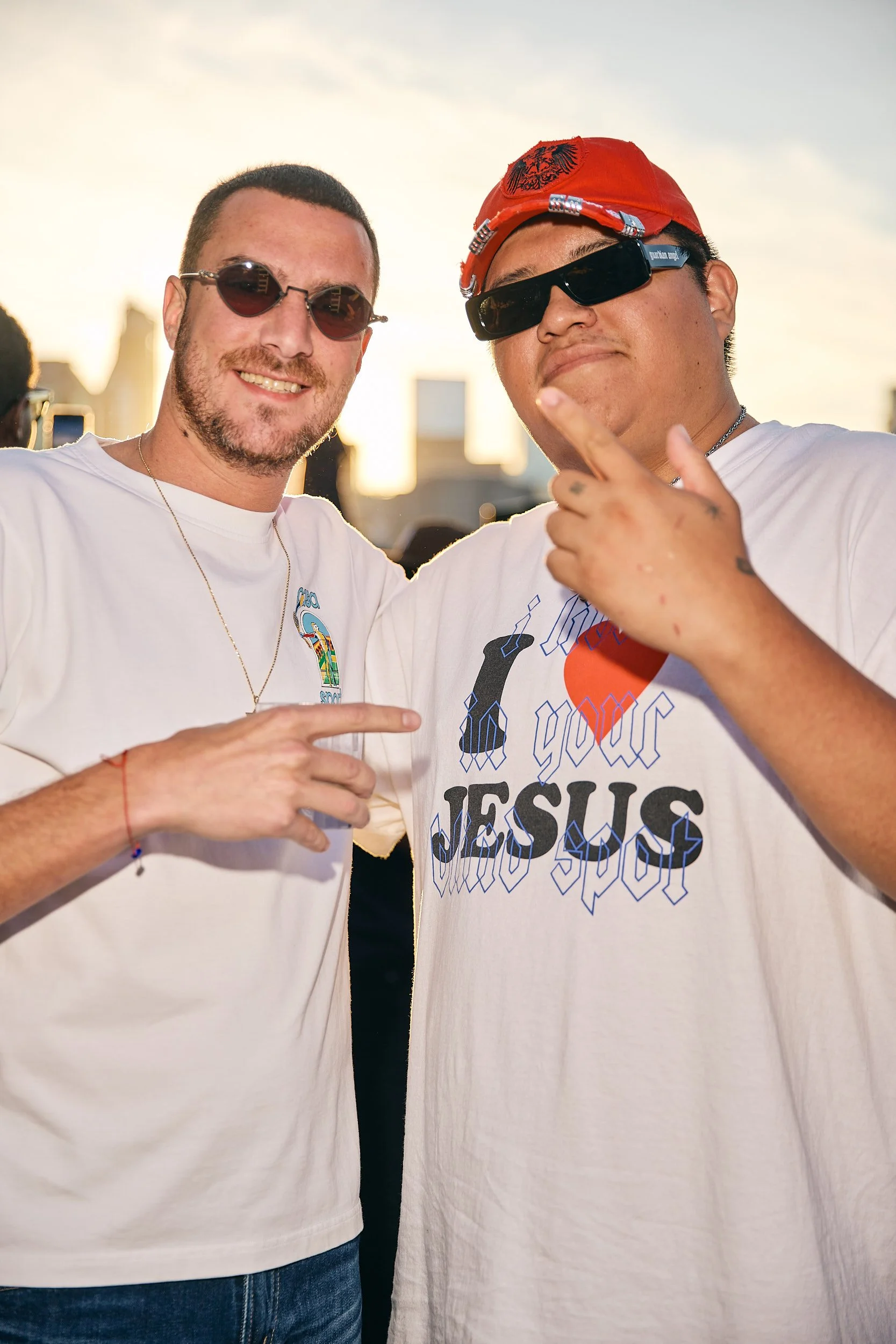 Two men standing together outdoors during sunset, smiling, wearing sunglasses and casual T-shirts, with one pointing at the other's shirt.