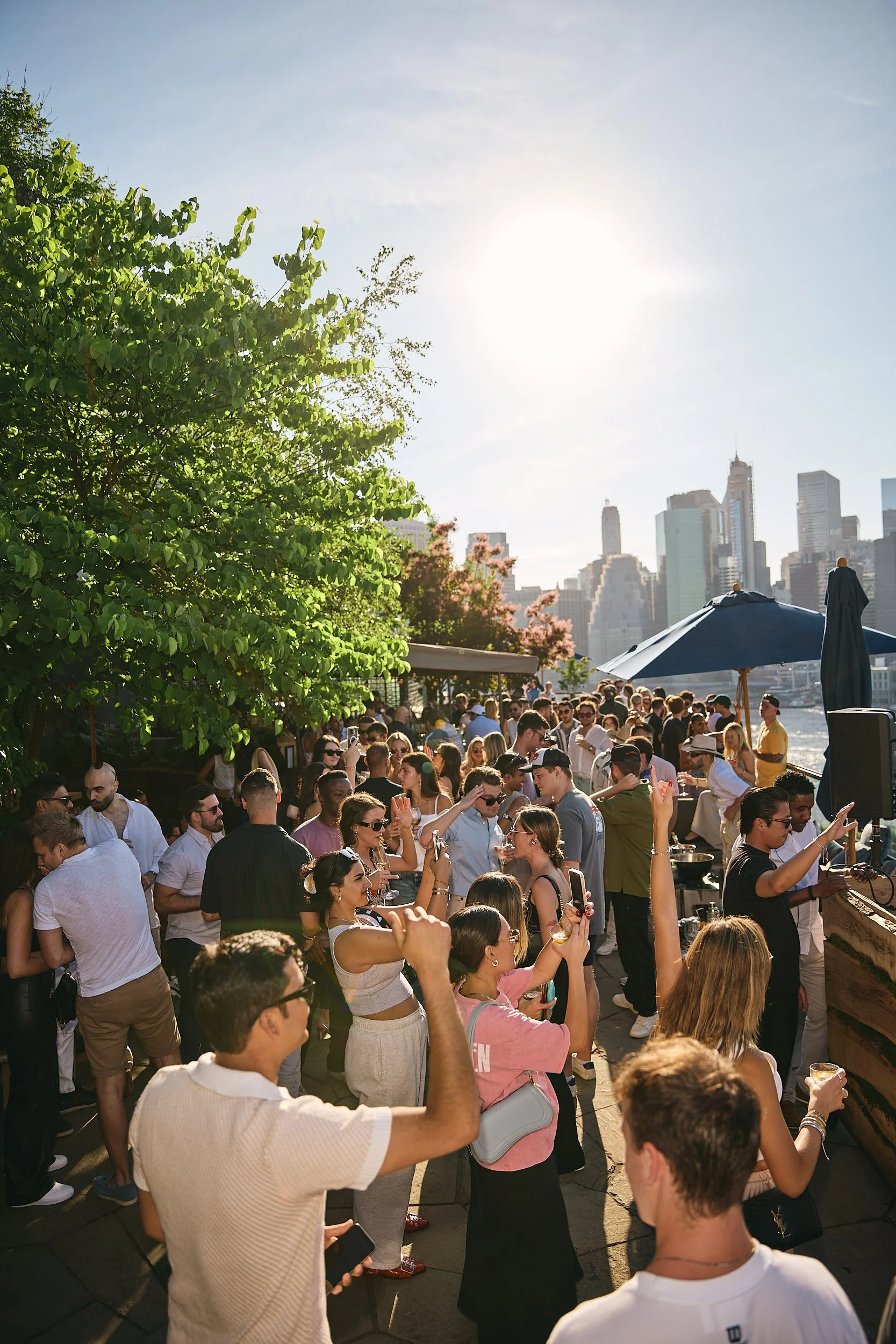 Crowd of people enjoying an outdoor event on a sunny day near the city skyline, with trees and umbrellas providing shade.