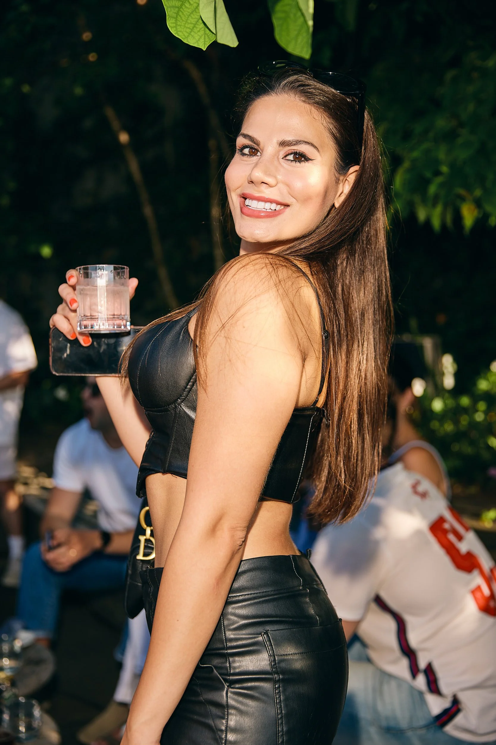 A woman with long brown hair wearing a black leather crop top and leather skirt, smiling and holding a glass of drink outdoors with greenery in the background.