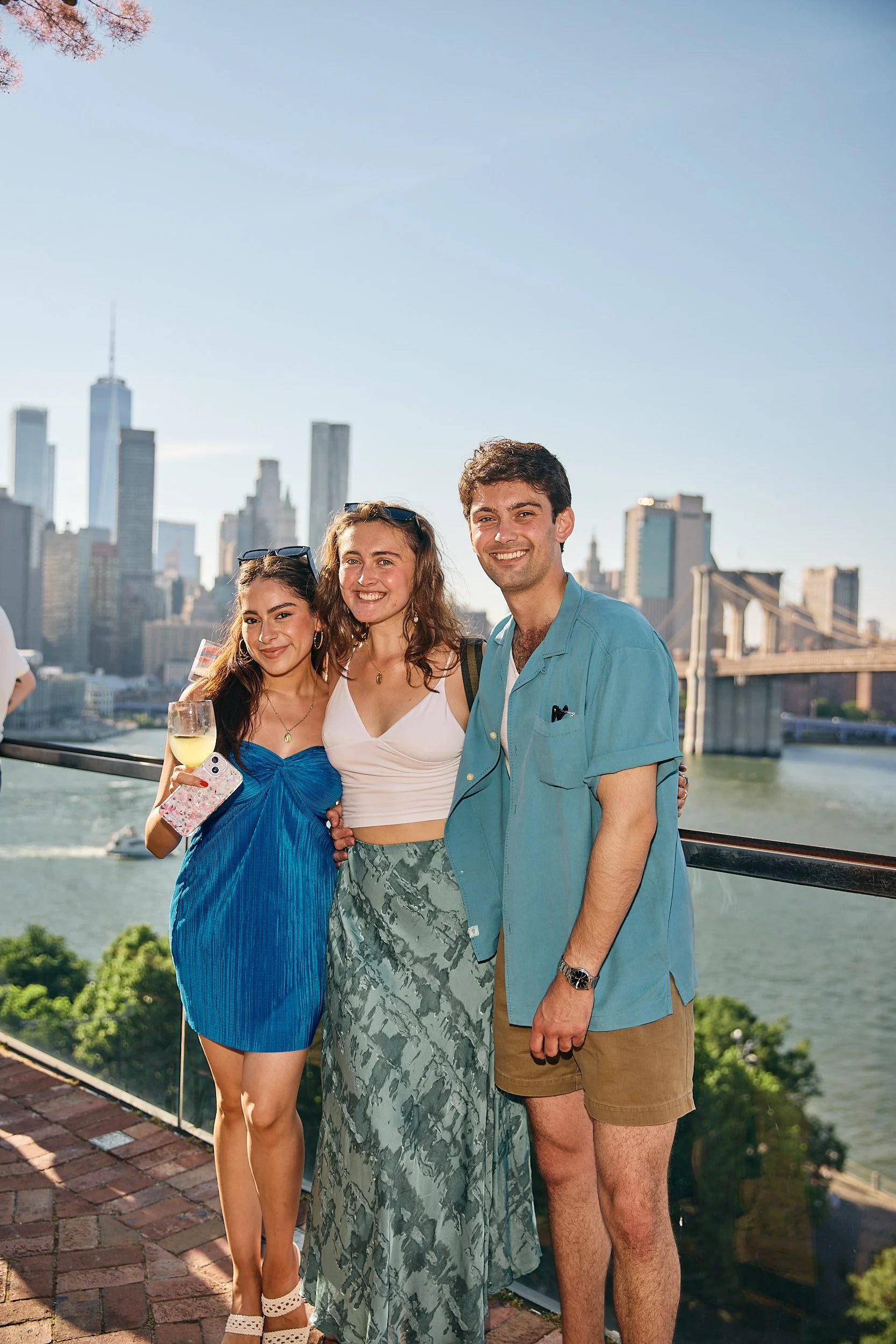 Three young adults smiling on a rooftop with New York City skyline and Brooklyn Bridge in the background, one holding a wine glass.