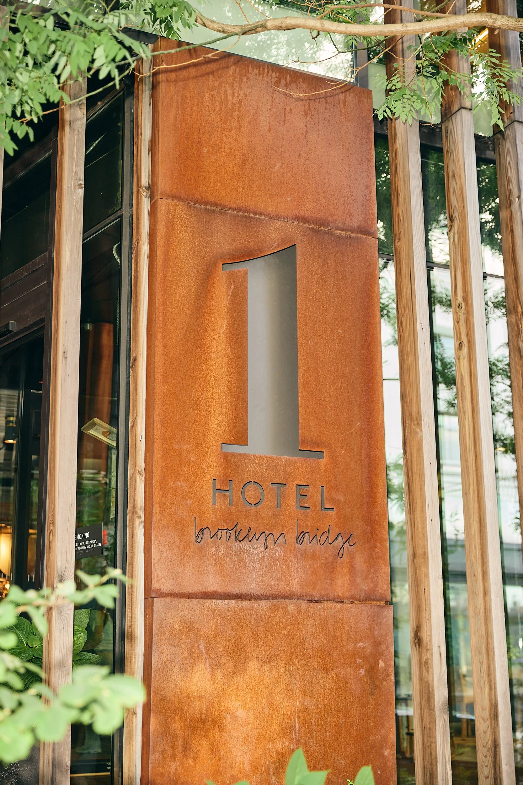 Exterior sign of Hotel Brooklyn Bridge with a large numeral 1 and rust-colored background, framed by wooden beams and greenery.