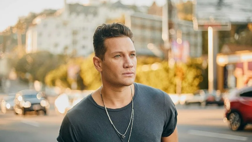 Young man with dark hair wearing a dark t-shirt and layered necklaces, standing outdoors in an urban setting during golden hour, with buildings and cars in the background.