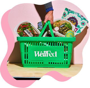 Green WellFed shopping basket filled with groceries on a wooden surface against a pink background.