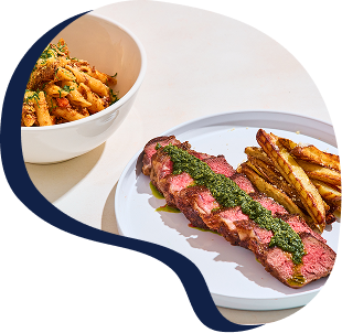 A plate with a sliced steak topped with green herb sauce and French fries, and a bowl of pasta salad with tomatoes and herbs in the background.