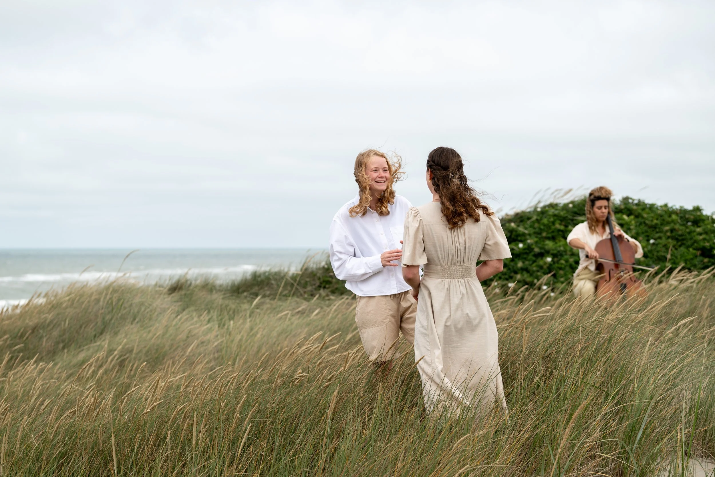 two girls standing close in the dunes with the ocean in the background. A woman playing cello in the background