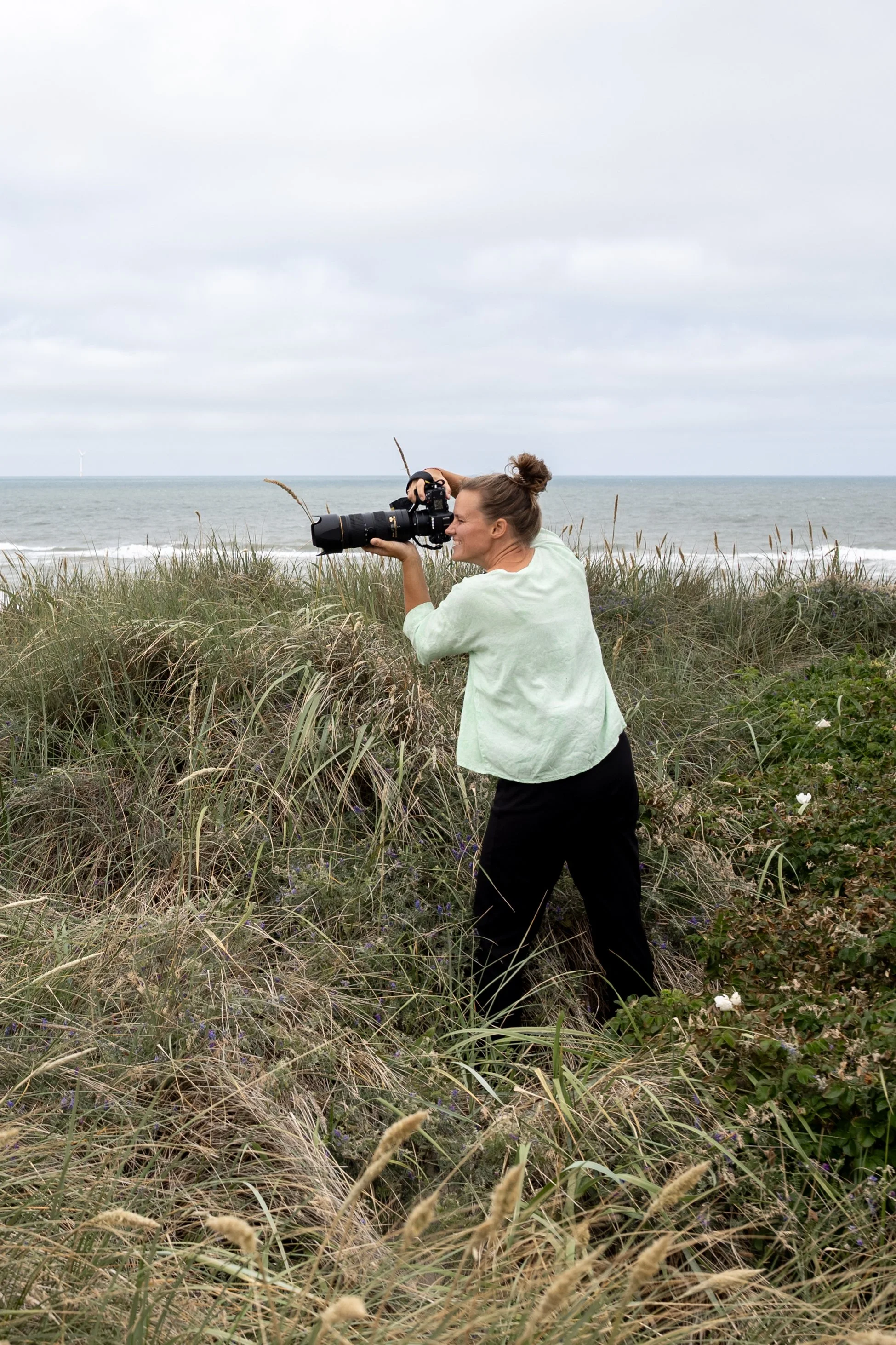 a woman standing with her big camera in the dunes near the ocean