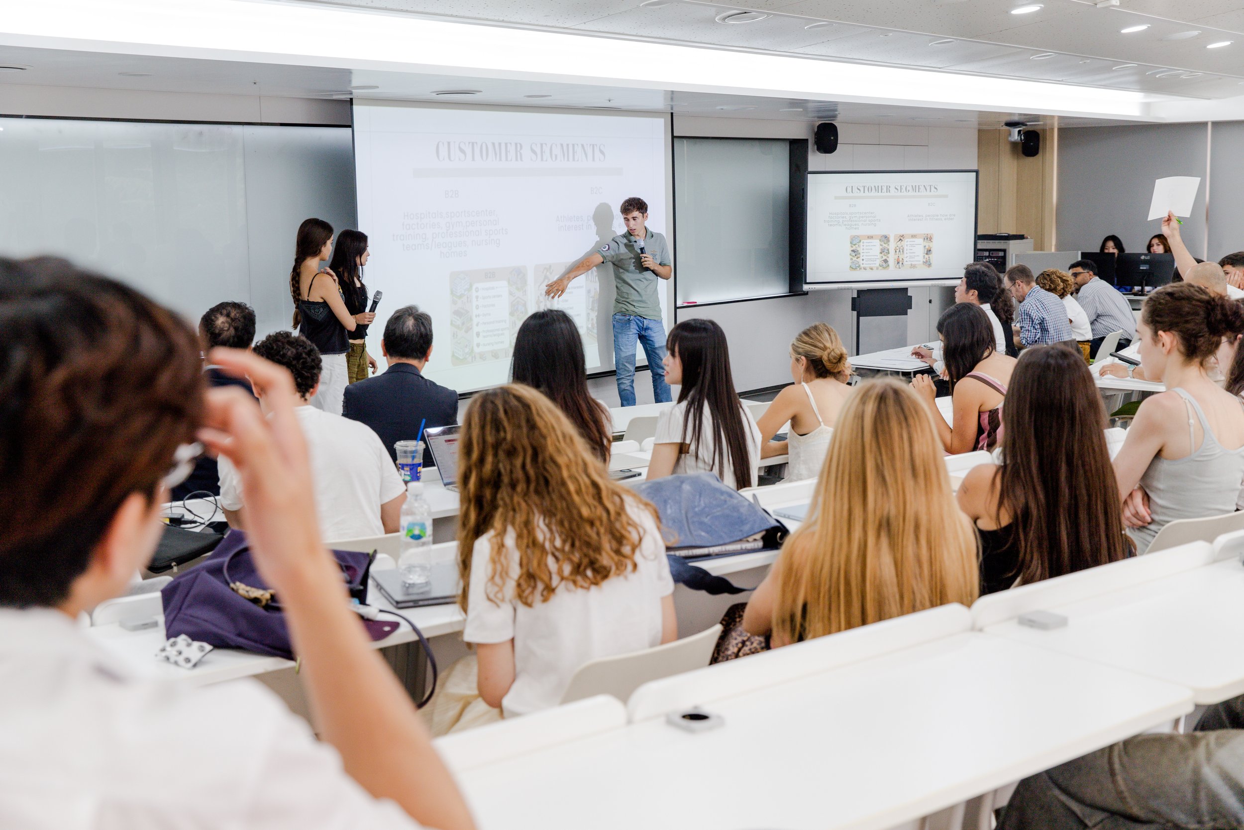 Una conferencia en un aula moderna con muchas personas sentadas y un presentador en el escenario señalando una pantalla con diapositivas sobre segmentos de clientes.