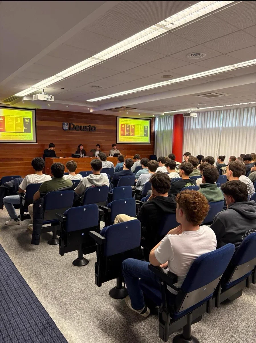 Grupo de estudiantes asistiendo a una conferencia en un aula con pantallas y en la pared un cartel que dice "Deusto".