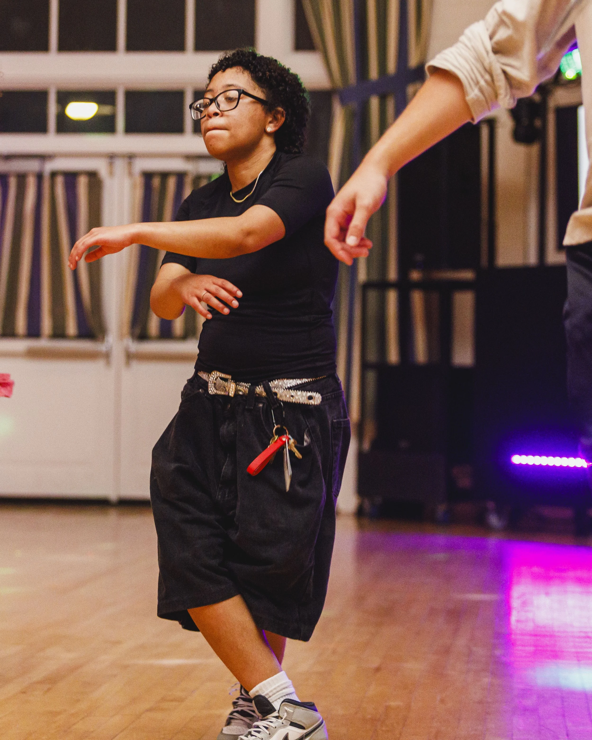 A young person with glasses and curly hair dancing on a wooden floor, wearing a black t-shirt, black shorts, a silver belt, and sneakers with white socks. They are in a room with windows and patterned curtains in the background, and there are colorfu