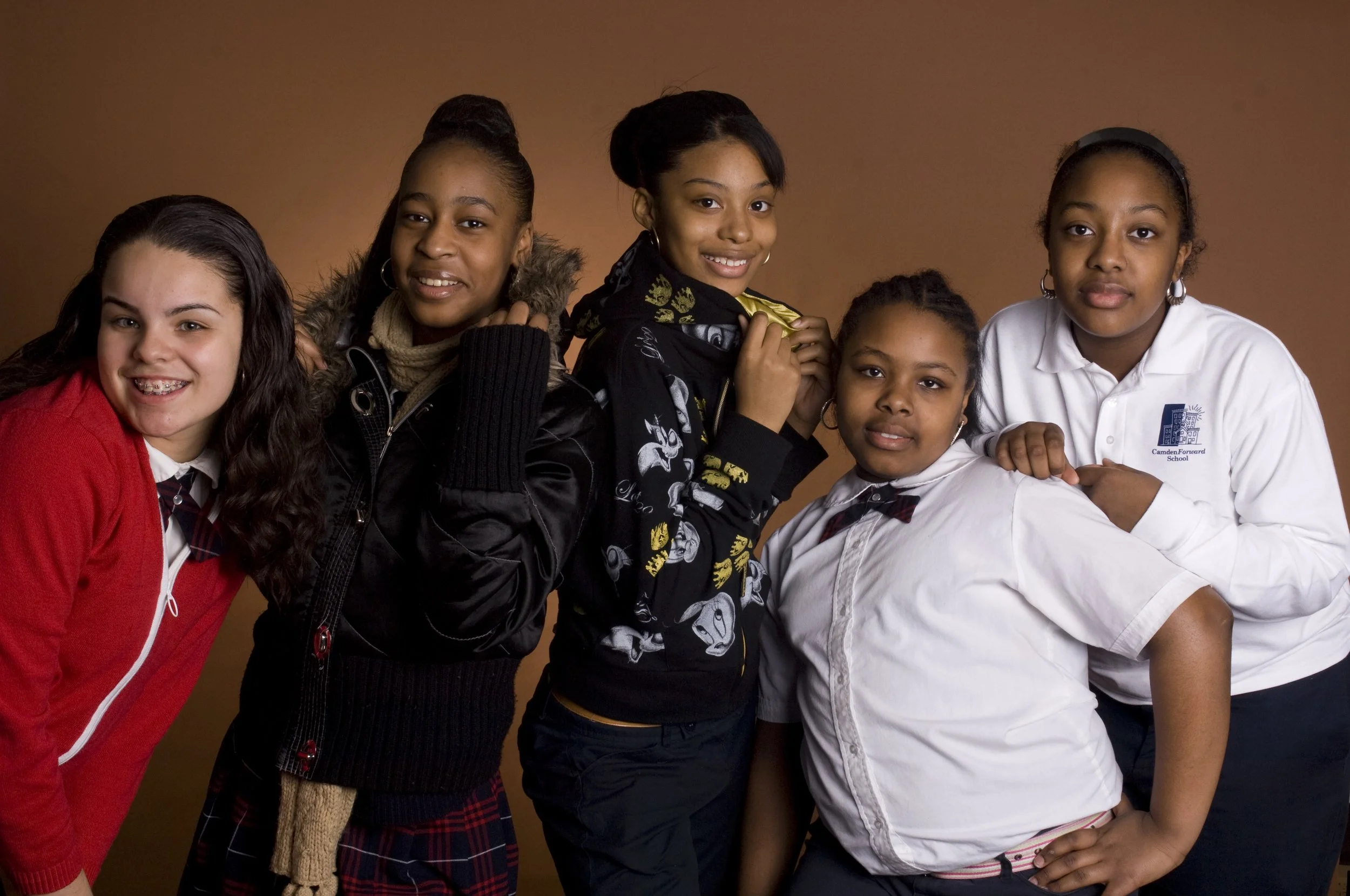Group of five diverse schoolgirls in school uniform and casual clothing posing together against a brown background.