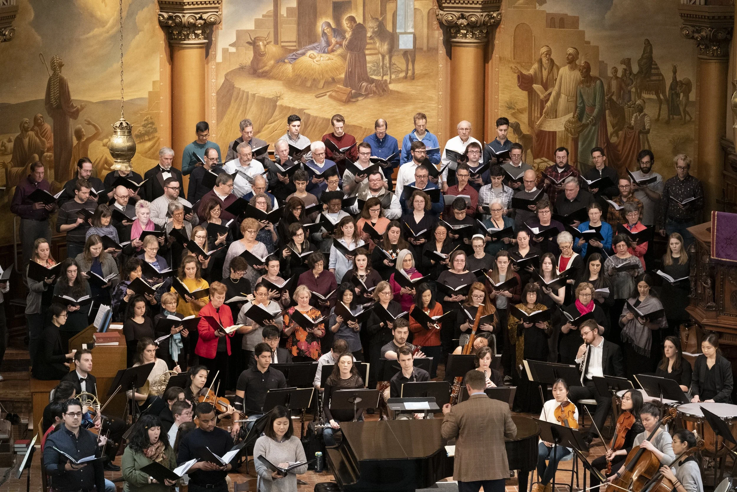 Large choir and orchestra performing in a church with ornate walls and a mural depicting biblical scenes.