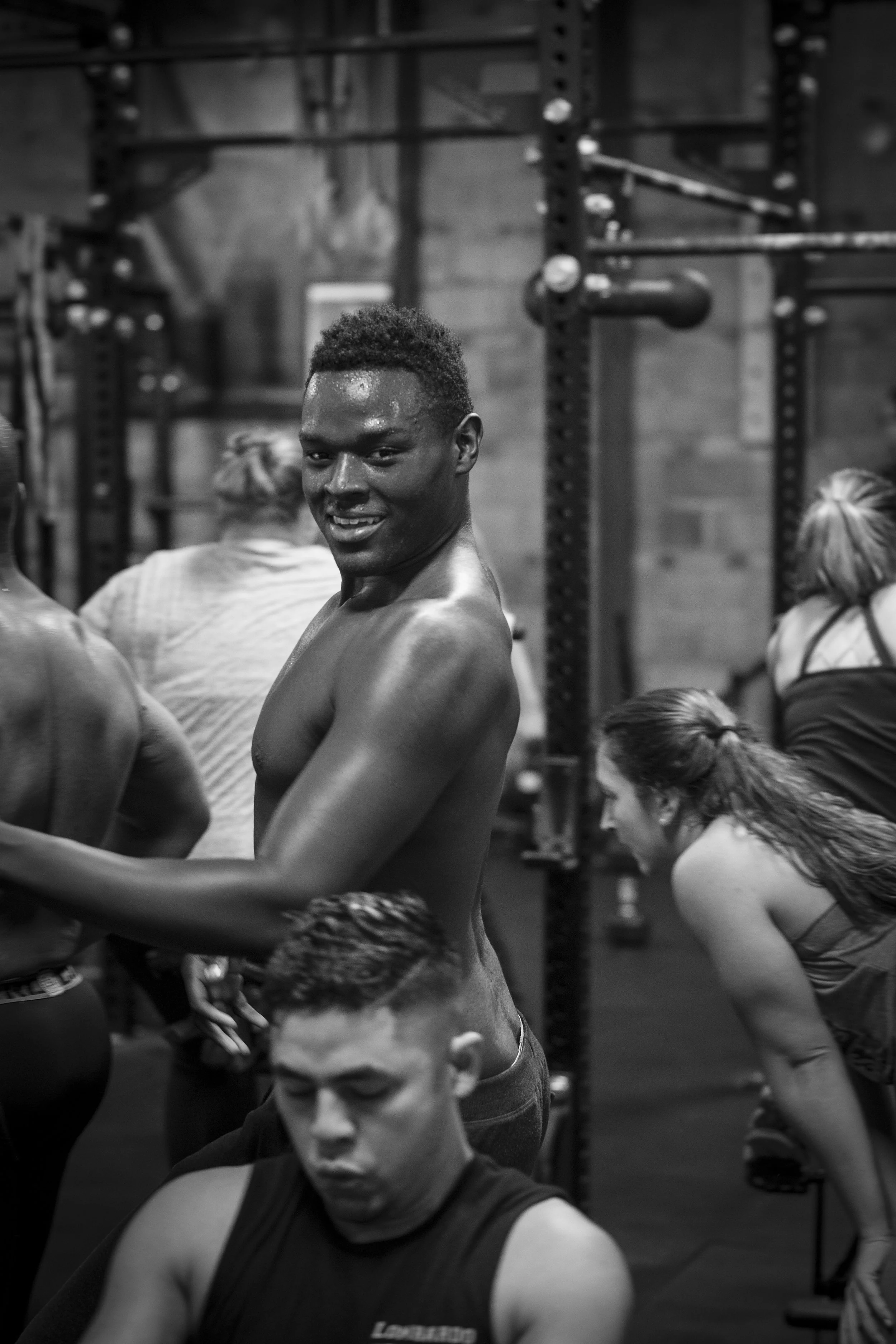 A shirtless man smiling at a gym, surrounded by other people exercising.