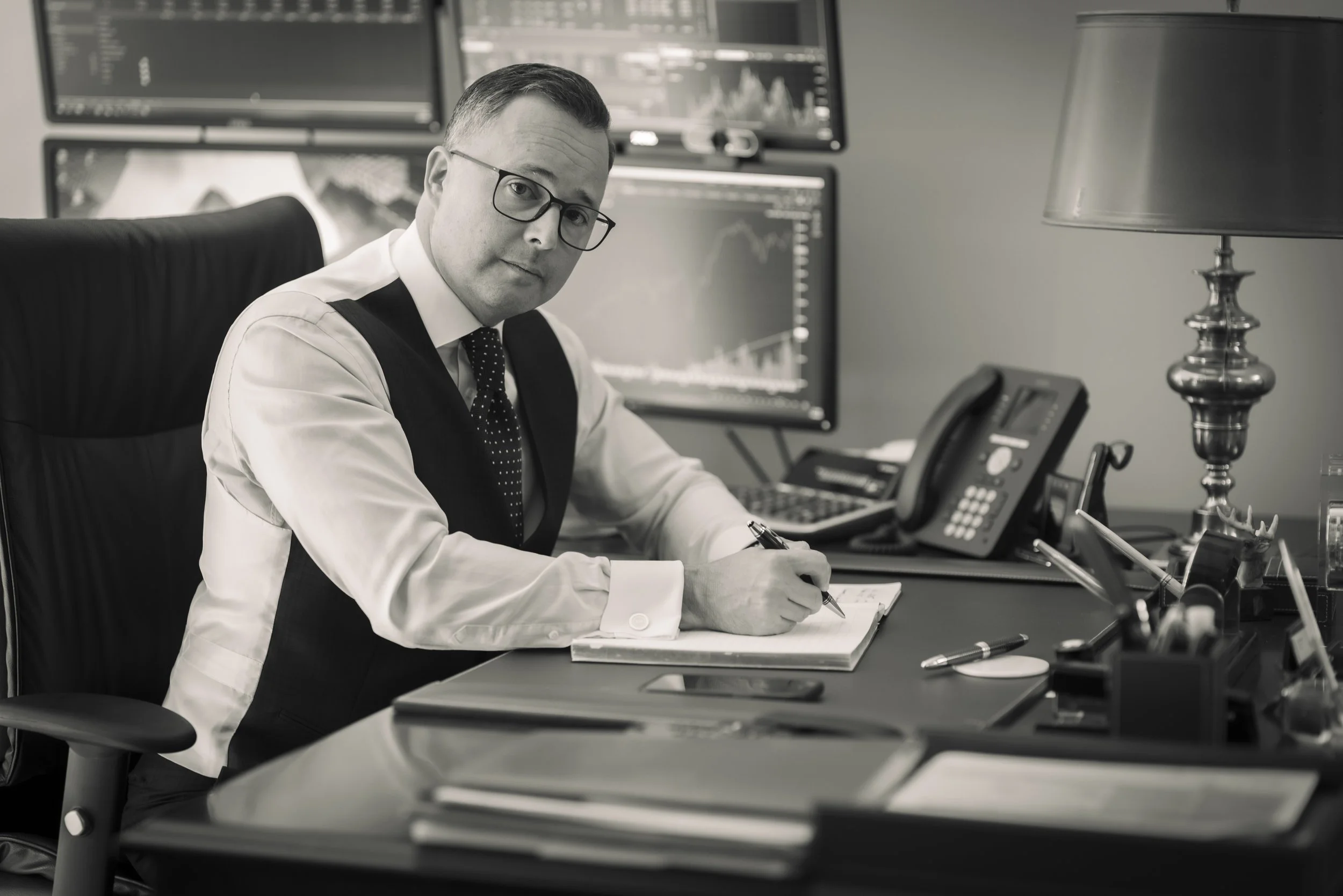 A man in a suit and glasses sitting at an office desk with multiple monitors displaying stock charts, writing in a notebook.