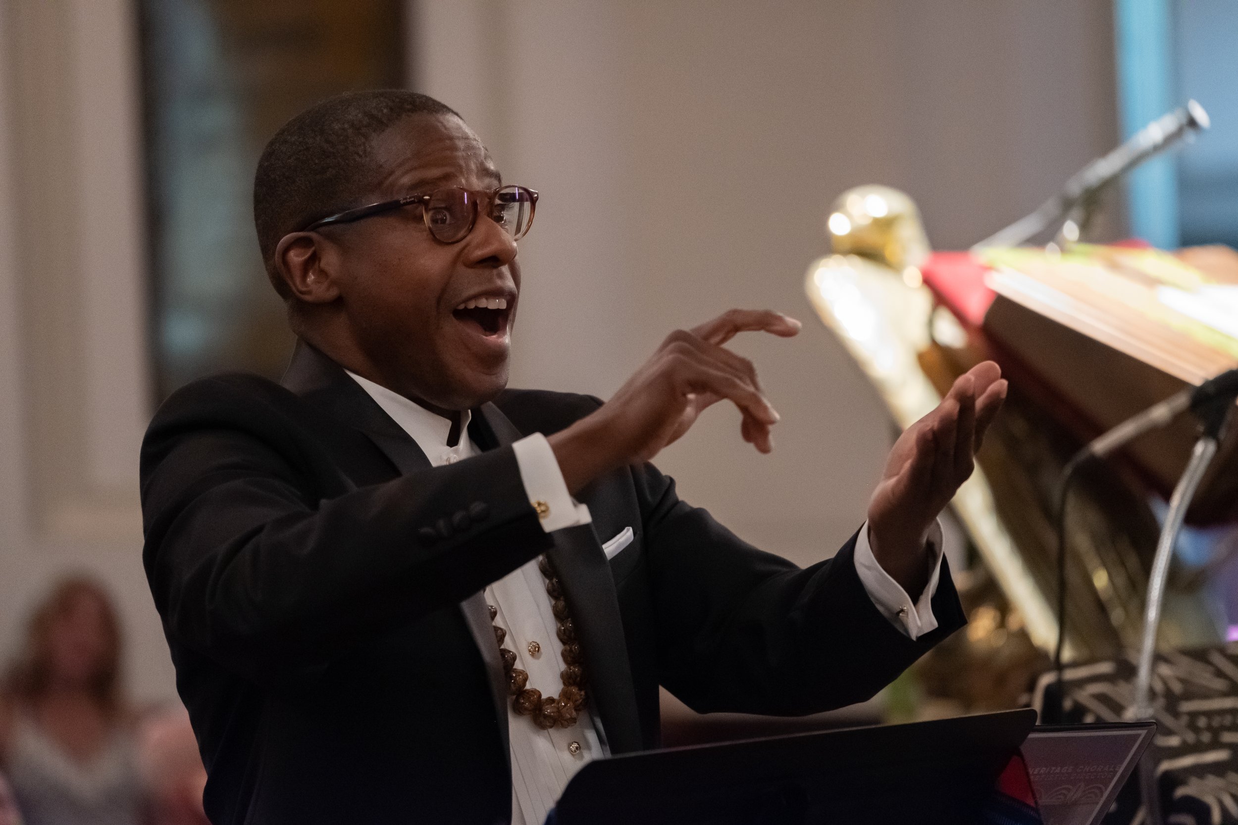 A man in formal attire, including a black suit, bow tie, and glasses, conducts the choir at a church or concert.