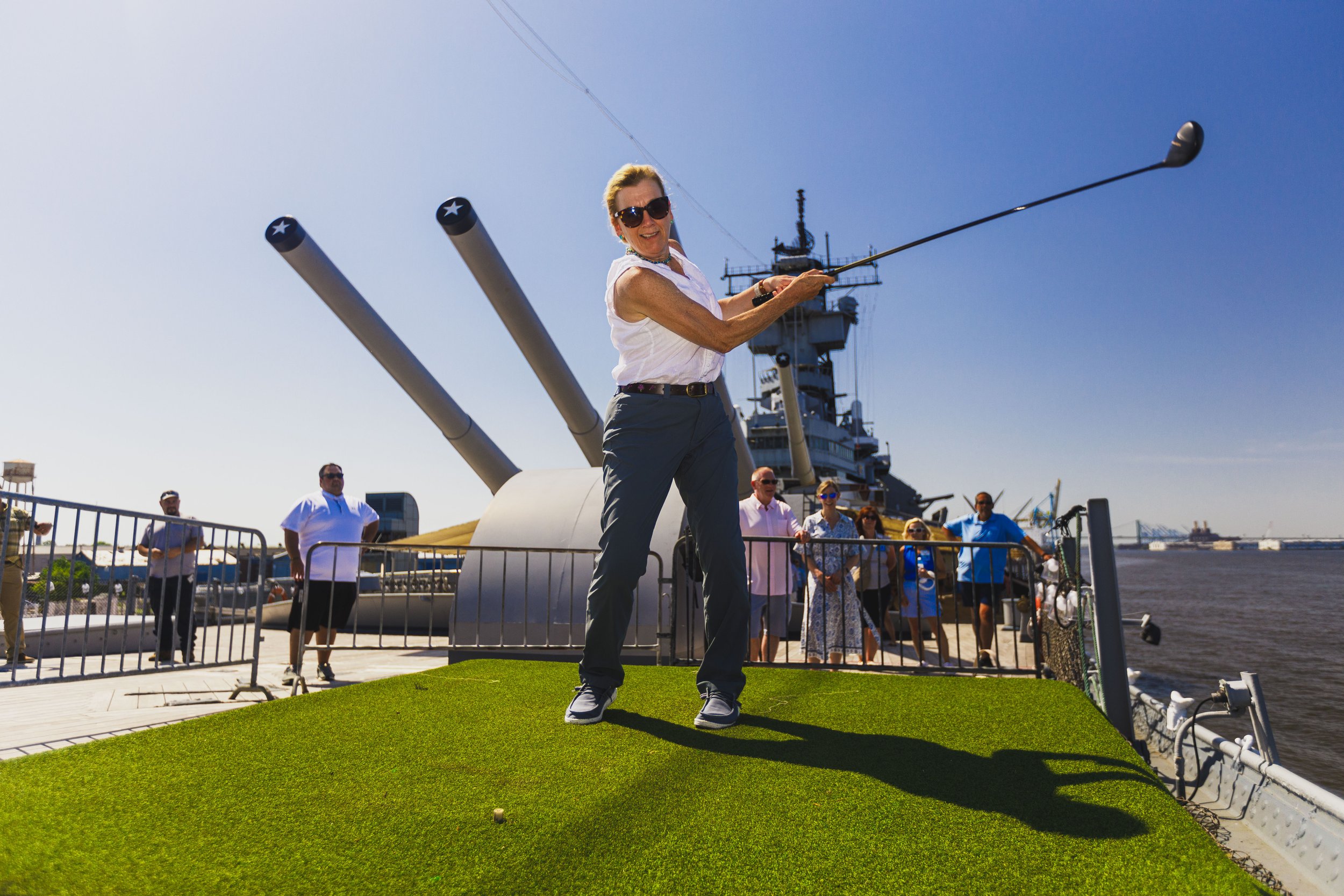 A woman playing mini golf on a green mat, with a battleship in the background and onlookers watching.