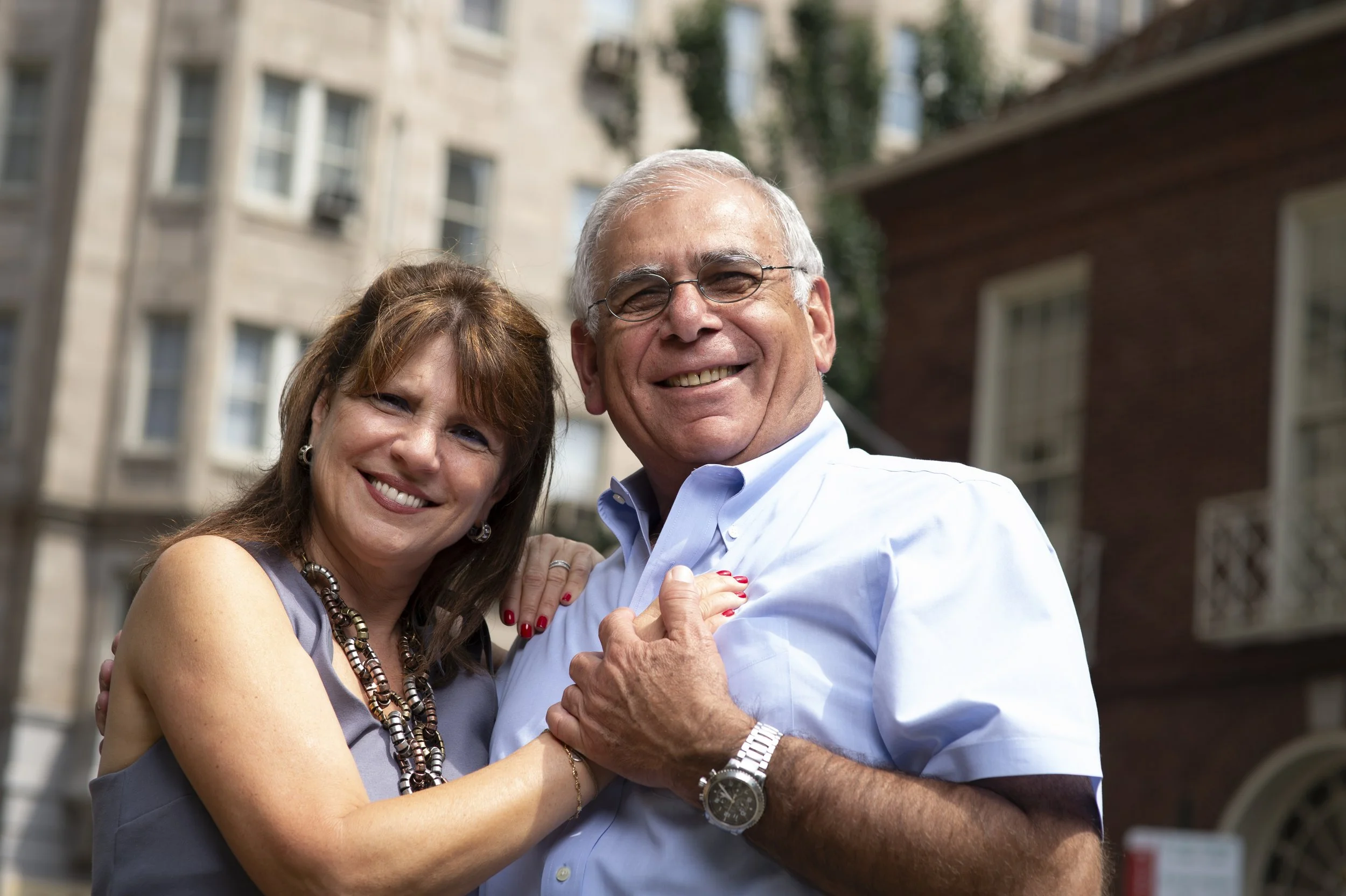 A middle-aged woman and man smiling and holding hands outdoors in a city neighborhood.