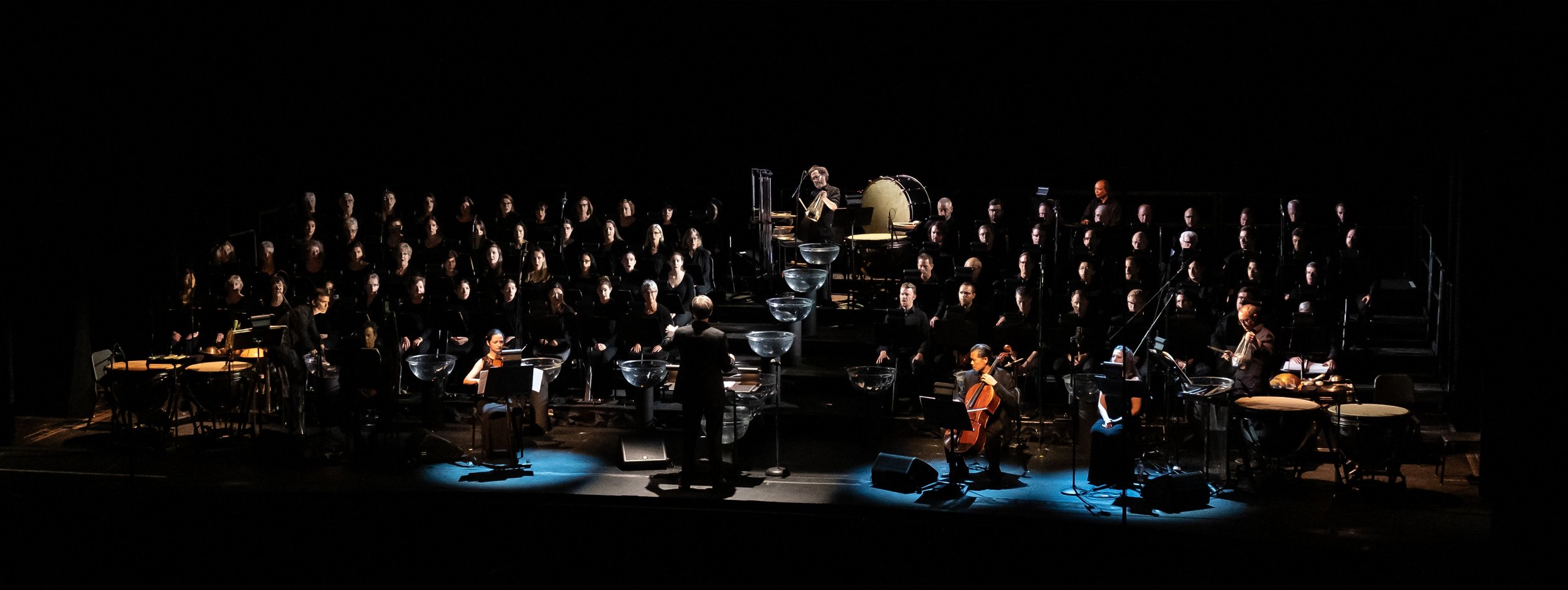 Orchestra performing on stage with conductor, string, percussion, and wind instruments, under dim lighting.