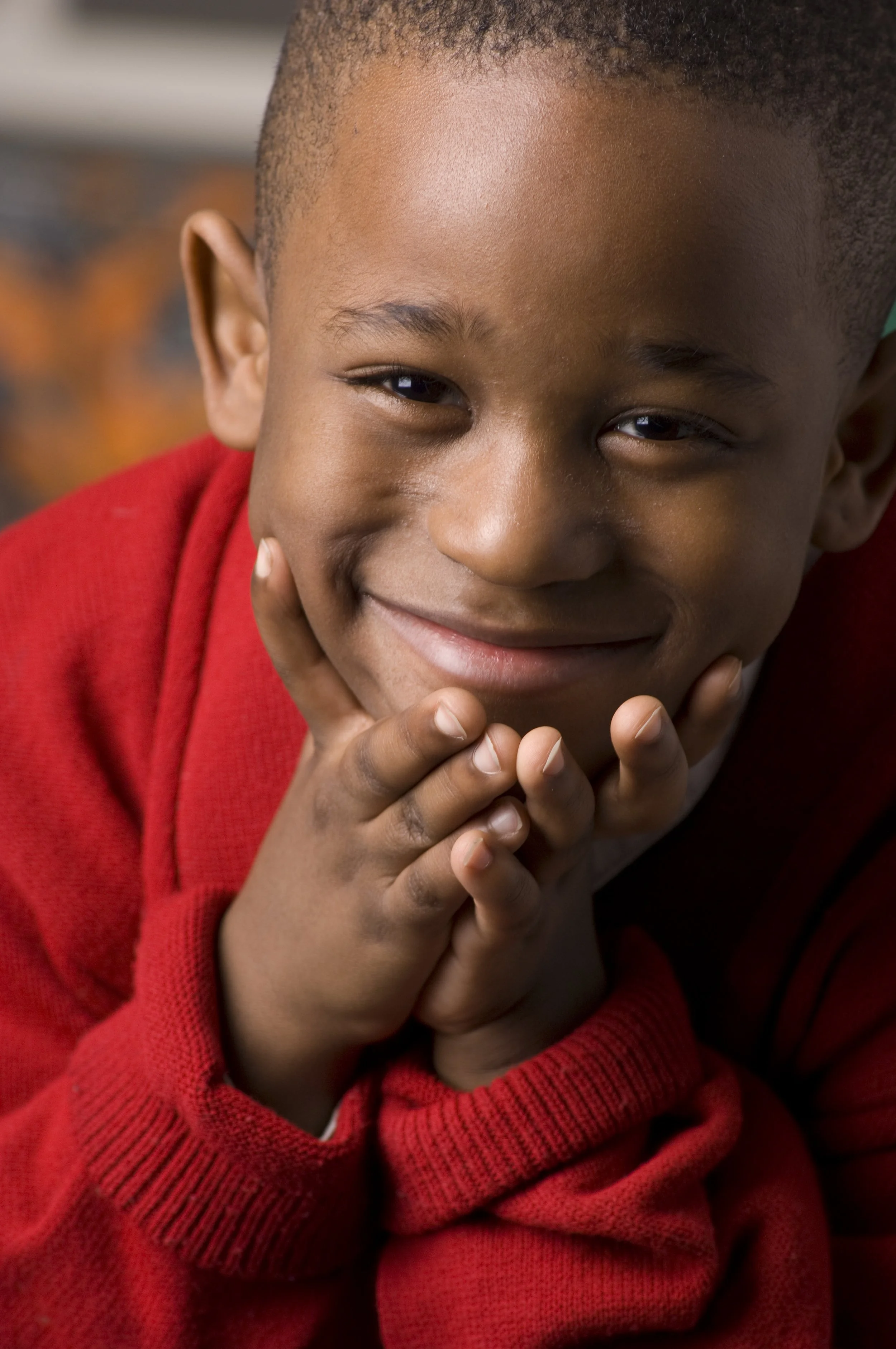 Close-up of a young boy smiling with his hands under his chin, wearing a red sweater.