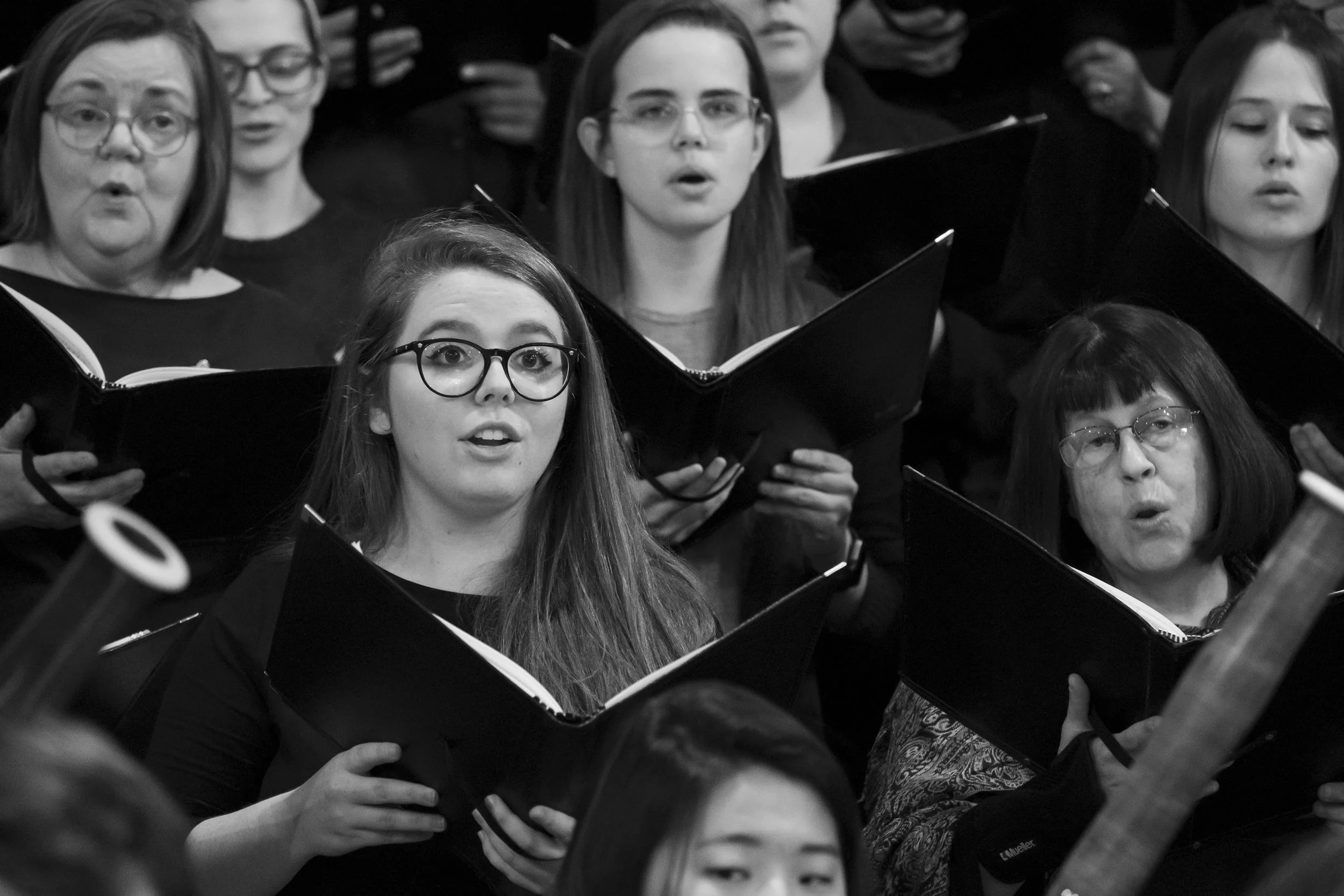 Black and white photo of a choir group singing, with women holding sheet music and appearing focused on their performance.
