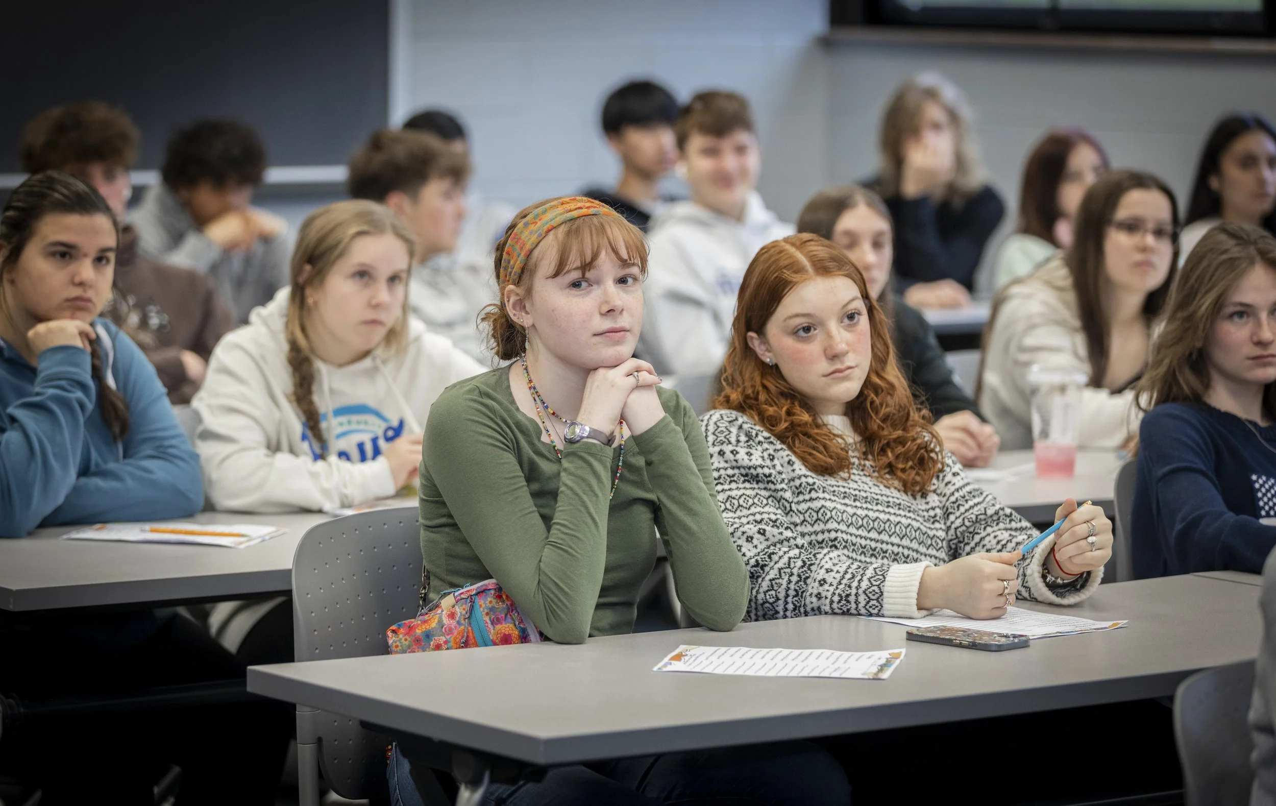 Classroom filled with students paying attention to a lecture, with some looking serious and focused.