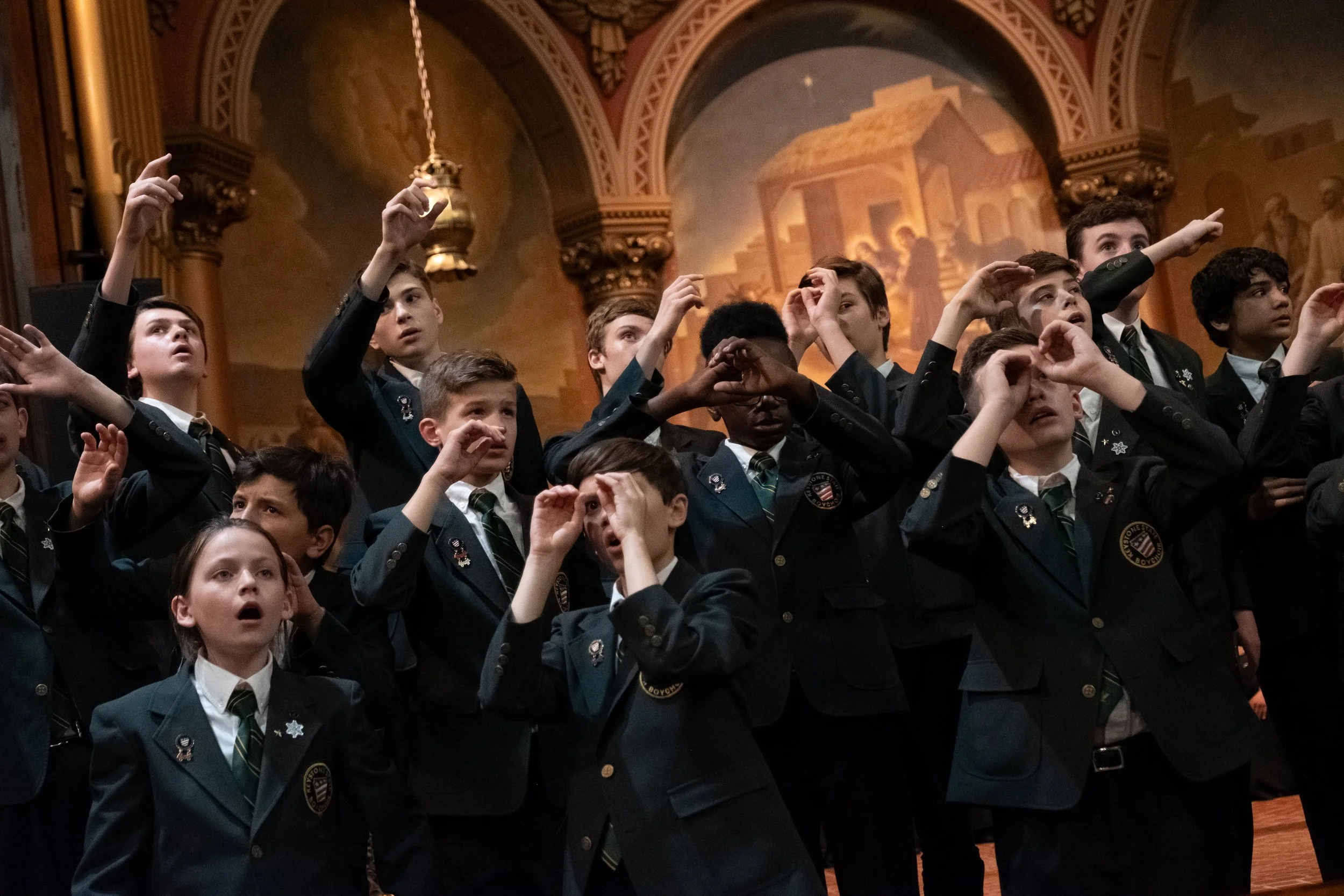 Group of boys in formal suits and ties raising their right hands in a salute or pledge inside a grand hall with ornate decorations and mural paintings on the walls.