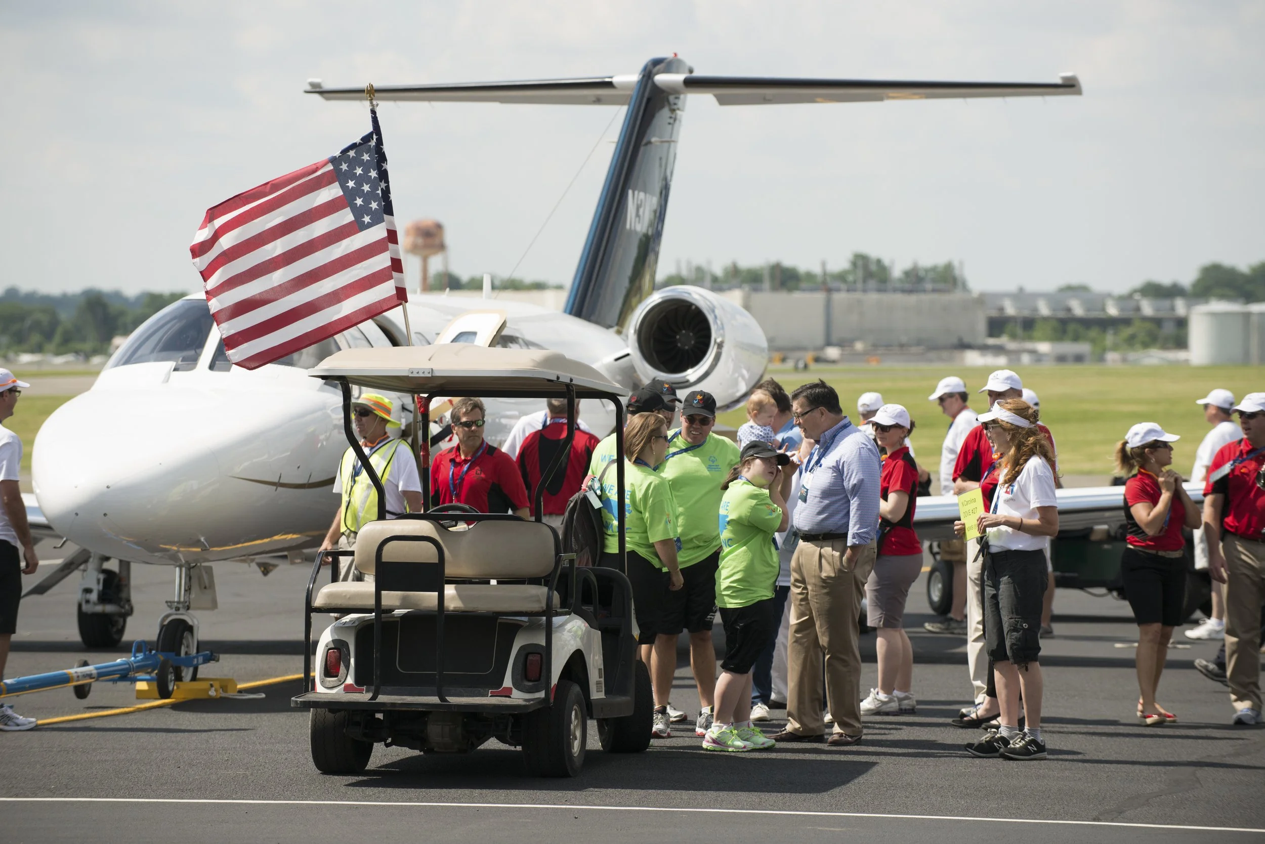 Group of people on an airport tarmac preparing to board a private jet, with an American flag attached to a golf cart in the foreground.