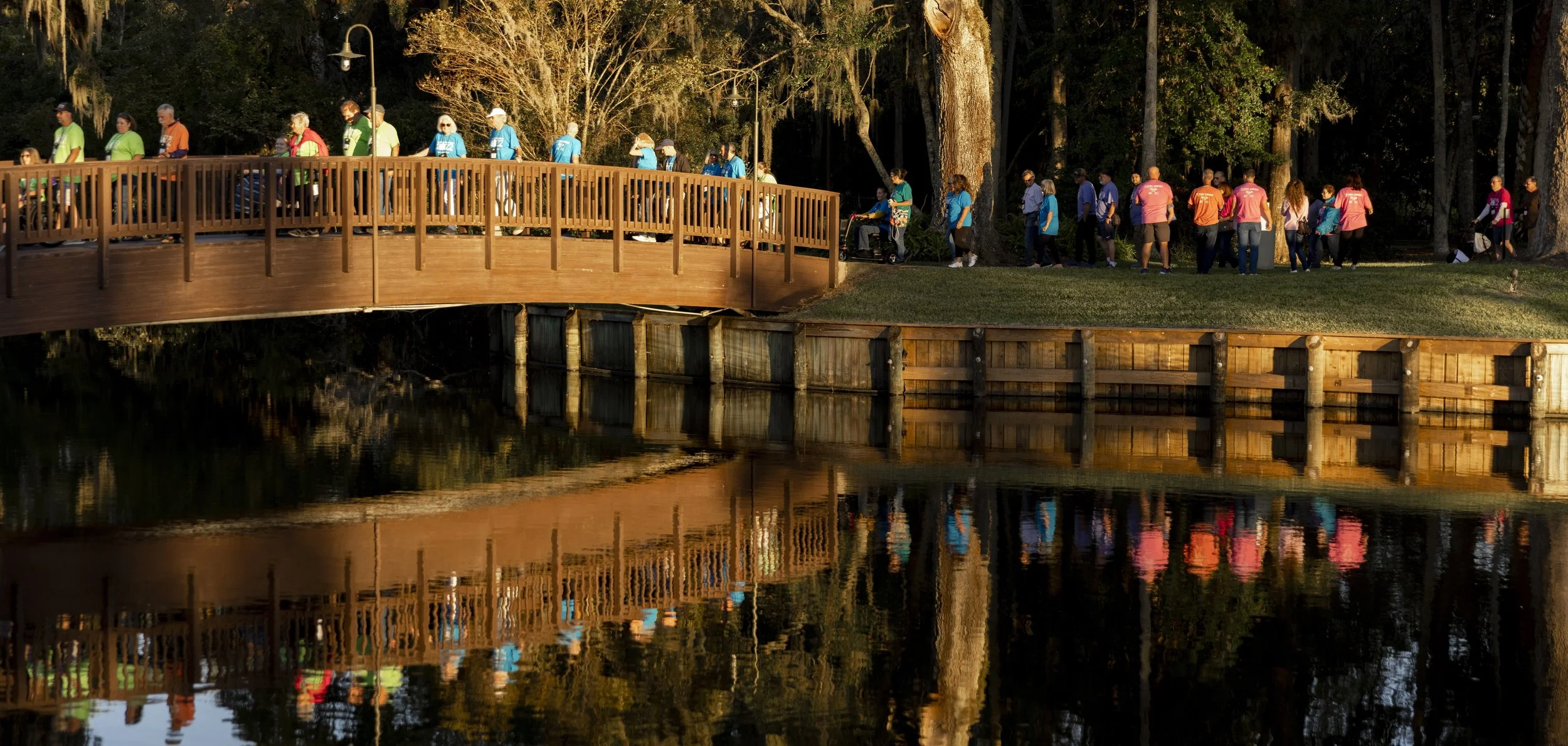 People walking in a line on a boardwalk over a body of water, reflecting the scene, with trees and outdoor lighting in the background.