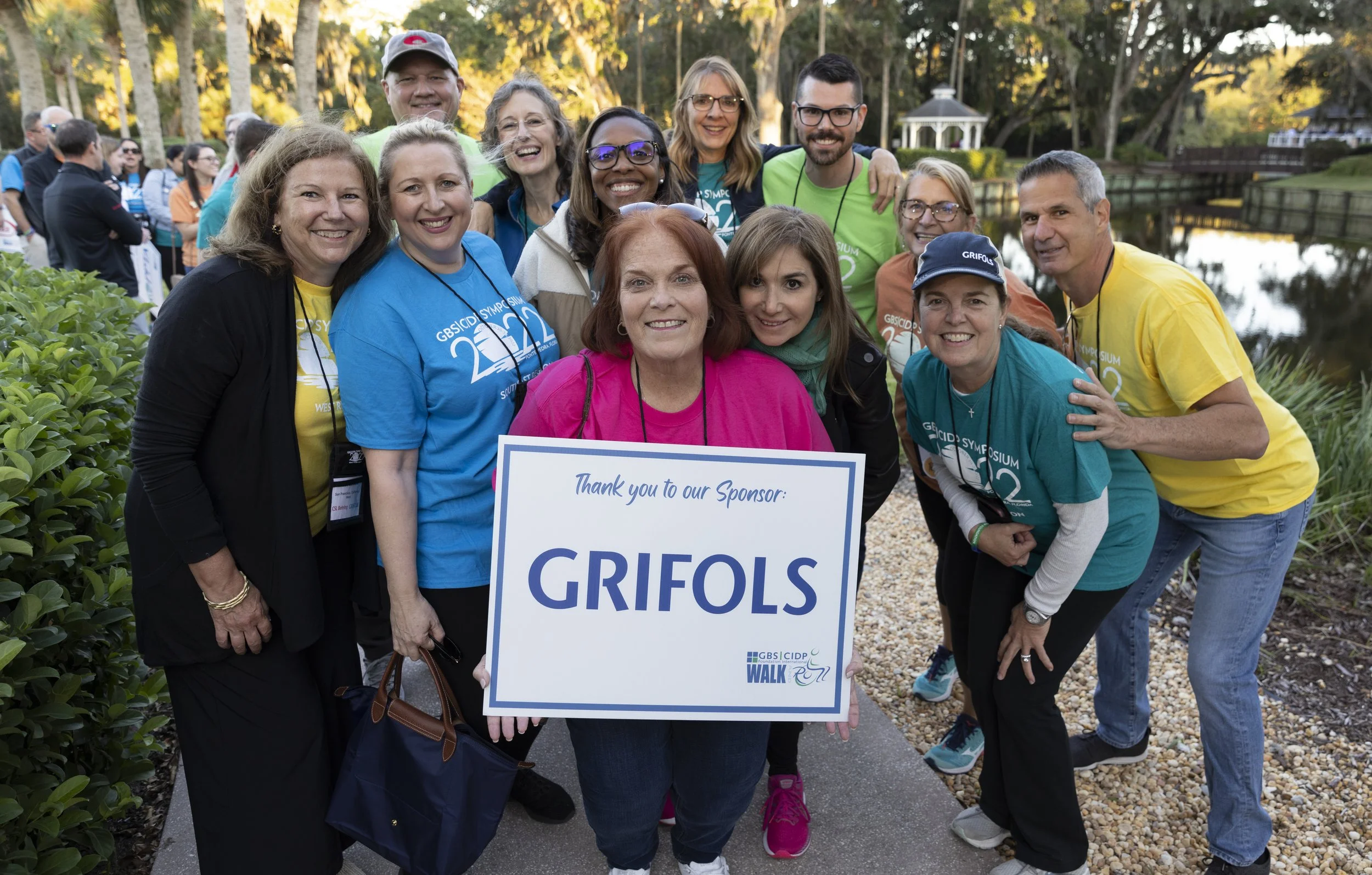 Group of diverse people smiling outdoors during a walk event, holding a sign that says 'Thank you to our Sponsor: GRI FOLS', with a pond and gazebo in the background.