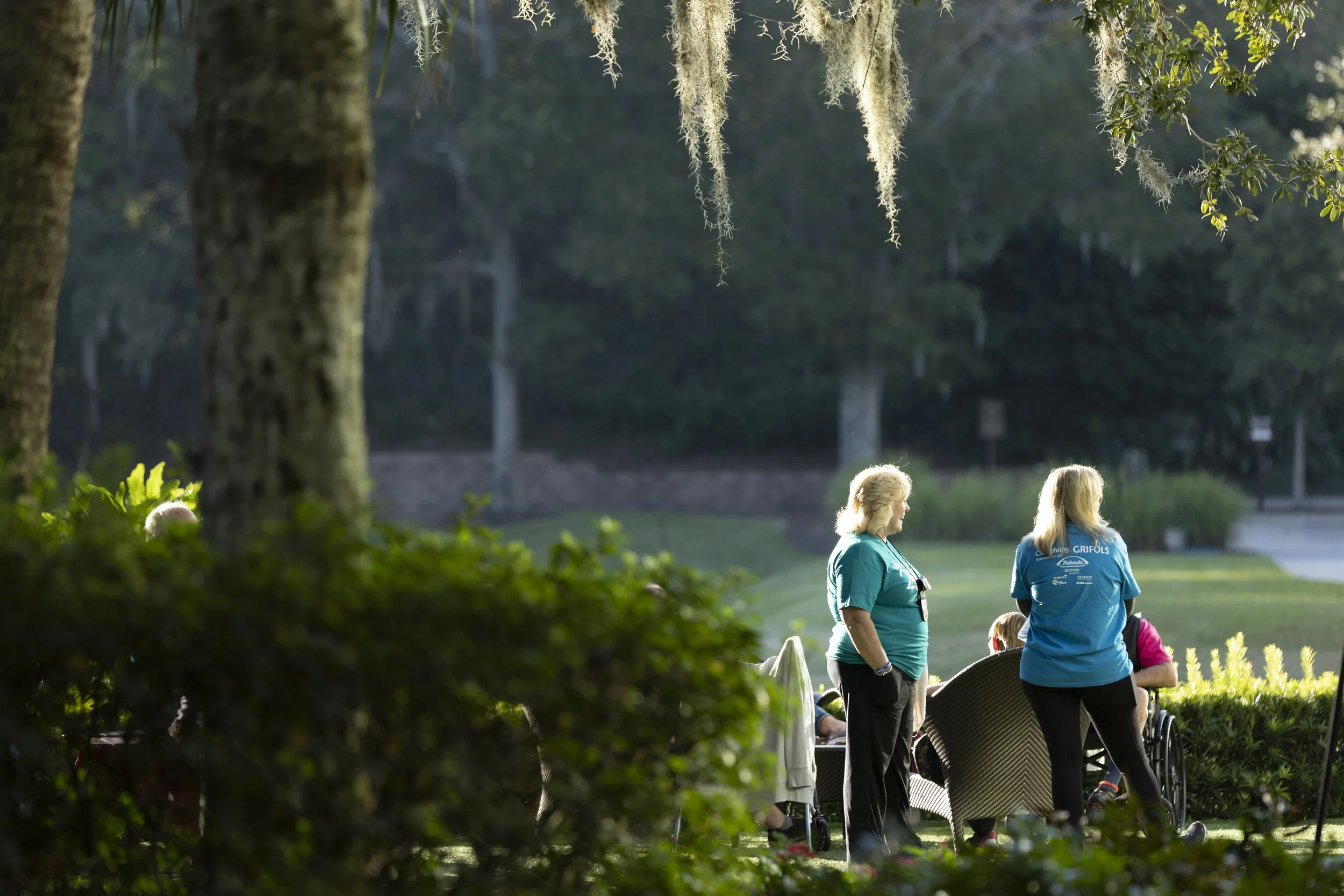 Group of people, including elderly women and a person in a wheelchair, gathered outdoors in a park during daylight, with trees and greenery around.