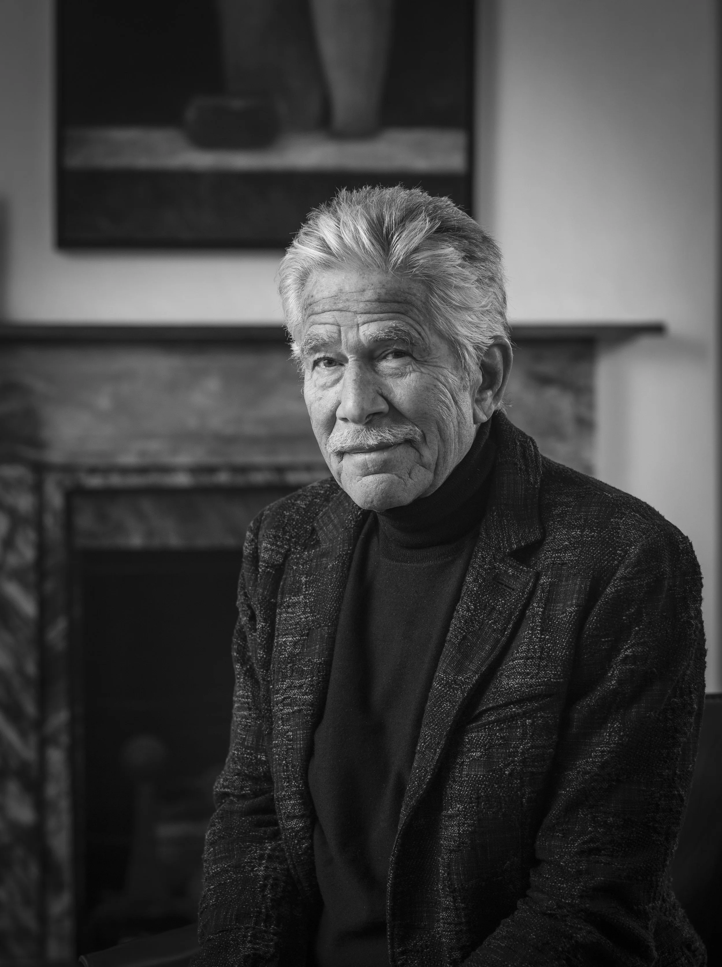 Older man with gray hair and a mustache, sitting indoors in front of a fireplace, wearing a dark turtleneck and a patterned blazer, looking at the camera.