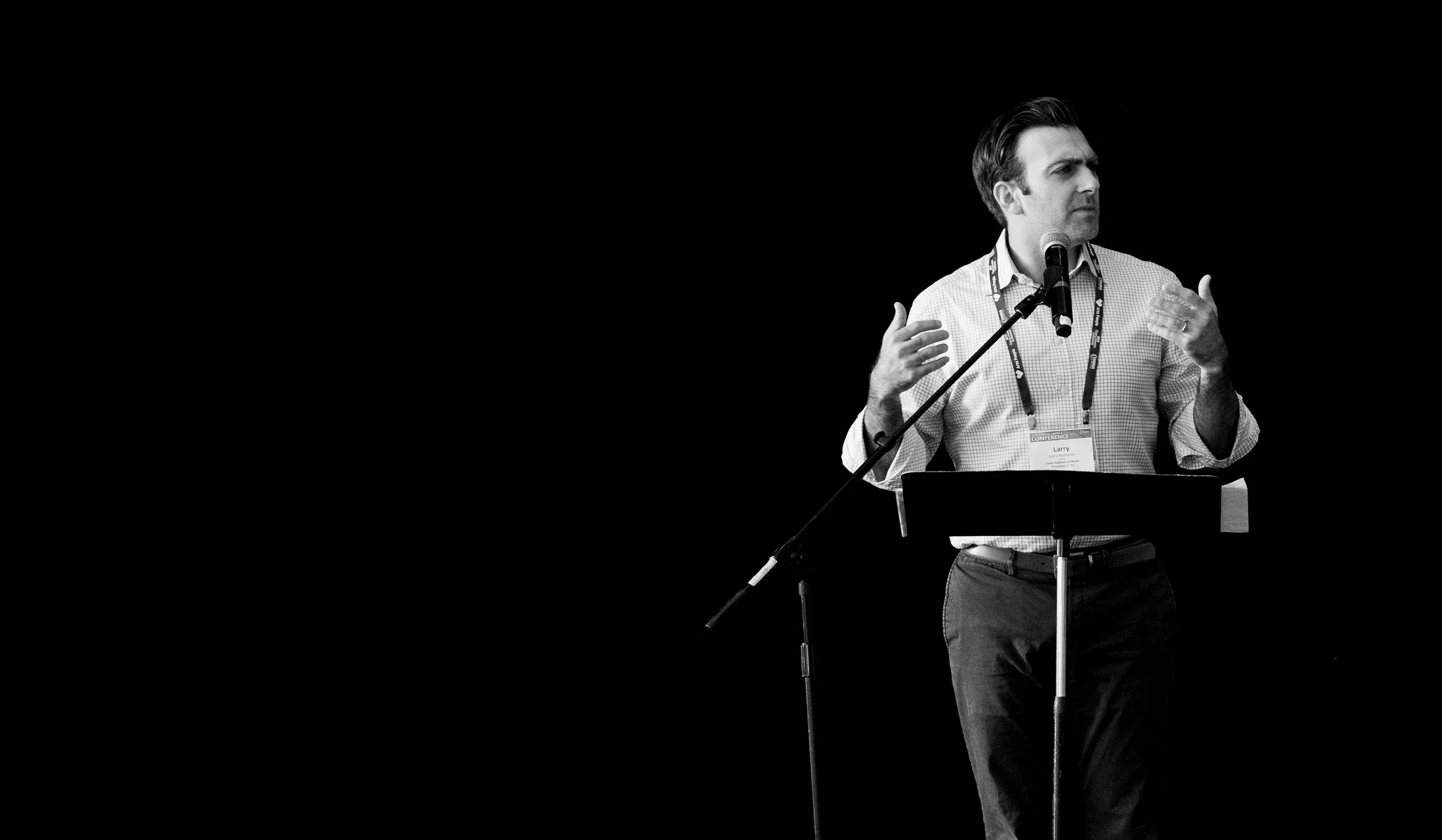 A man speaking at a podium with a microphone, gesturing with his hands, set against a black background.