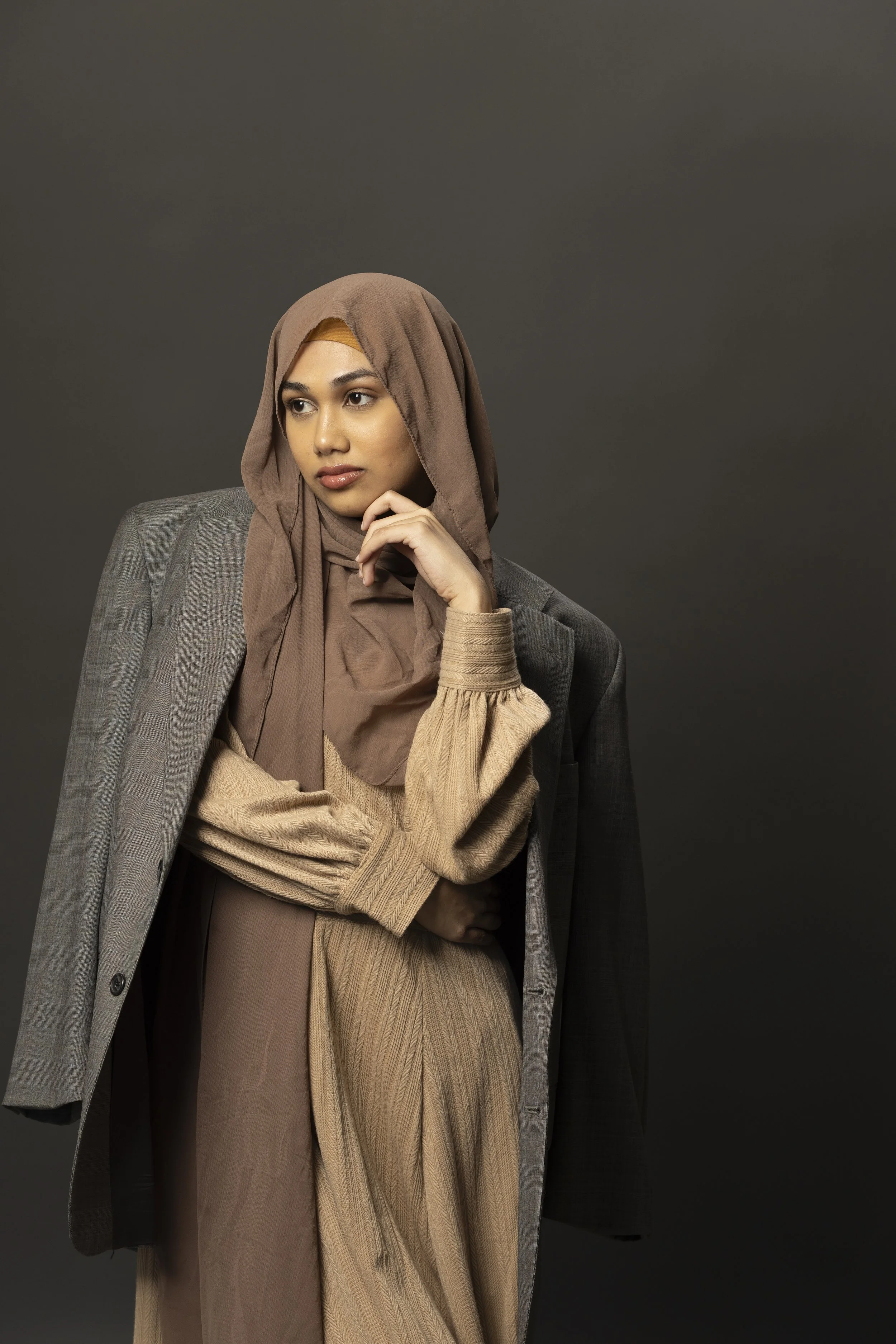 A woman wearing a taupe hijab, a beige and brown dress, and a gray blazer, posing against a dark gray background.