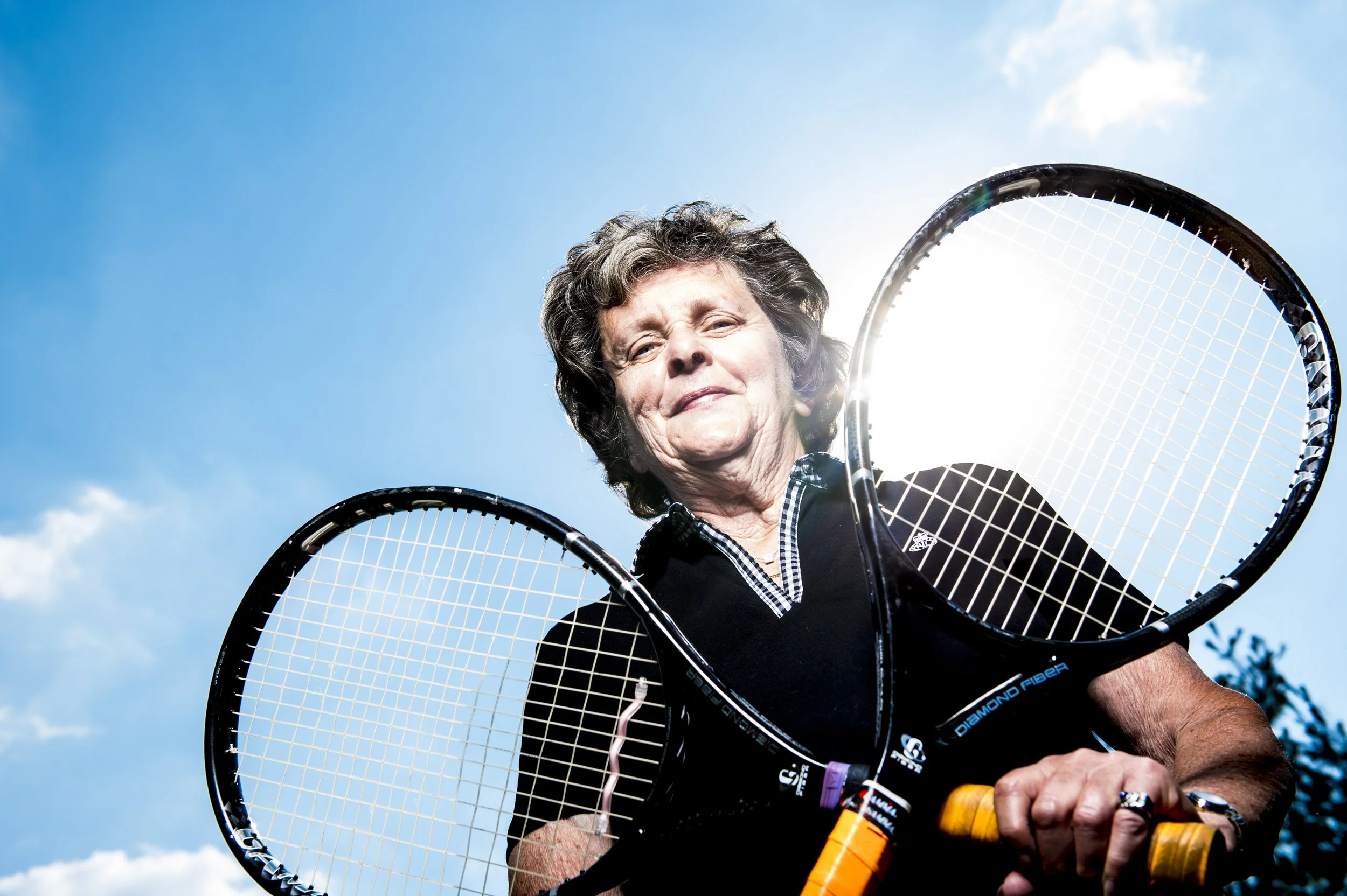Older woman outdoors holding two tennis rackets with a blue sky and sunlight in the background.