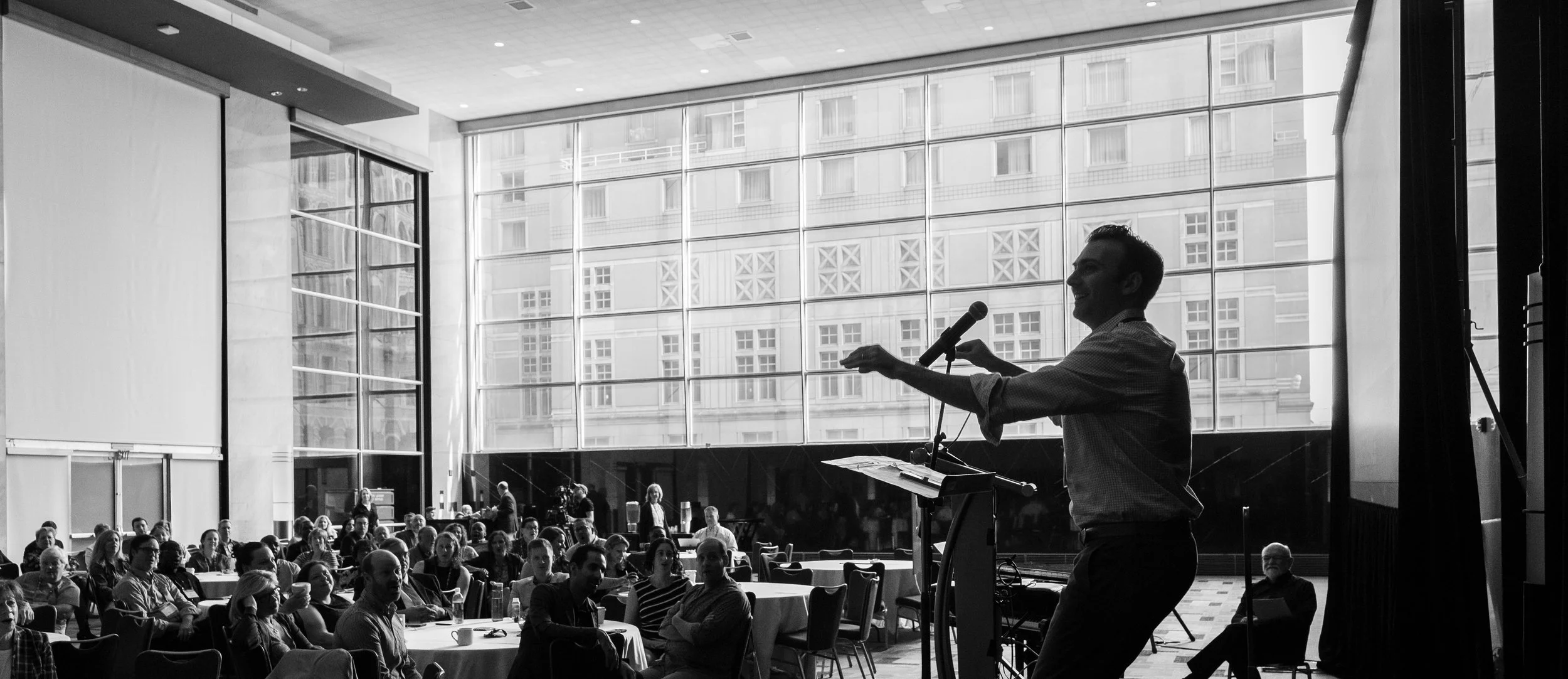 A speaker presenting at a conference with an audience in a modern, glass-walled room.