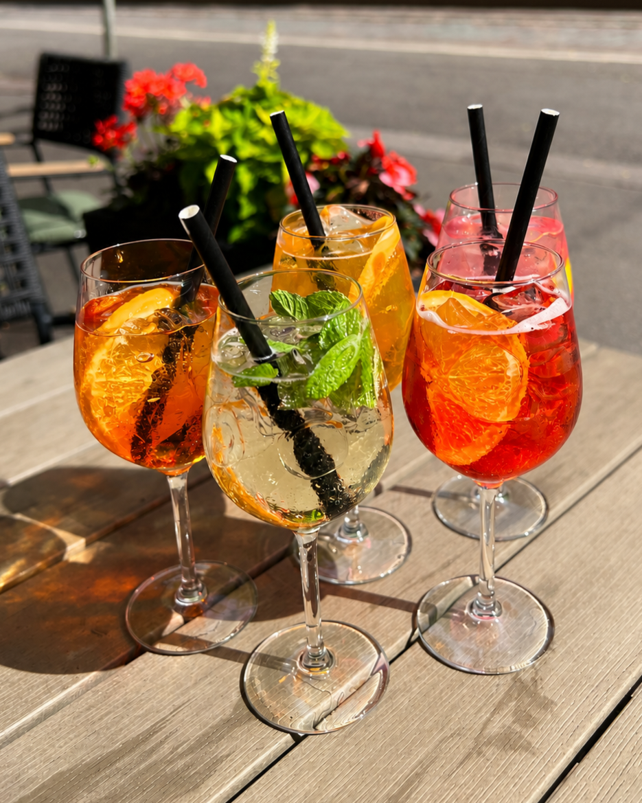 Neljä erilaista spritz-drinkkilasia, jotka sisältävät appelsiineja ja limenlehtiä, asetettuna puiselle terassipöydälle auringonpaisteessa, taustalla kukkapenkki ja kaupungin katu.