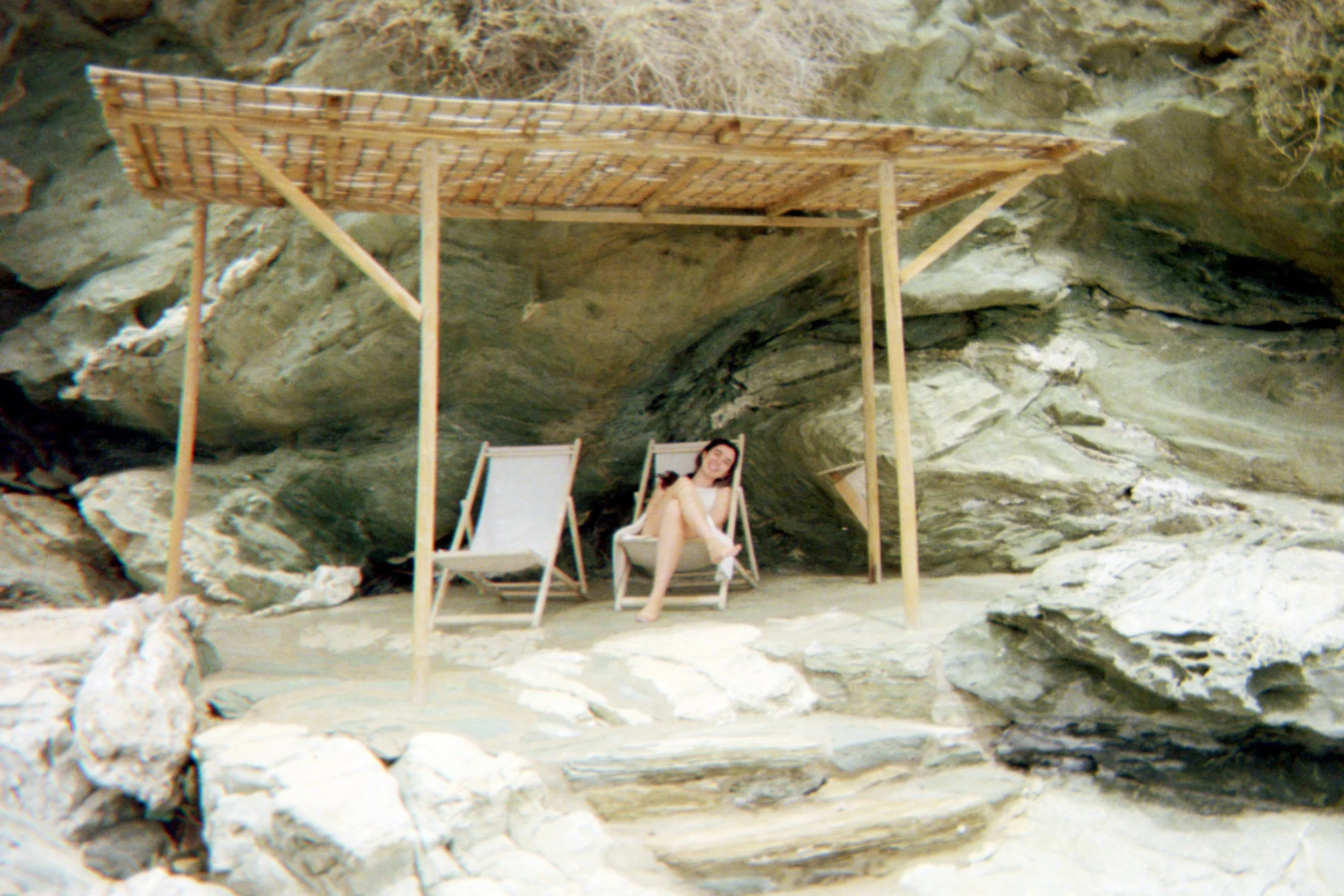 Person relaxing on deck chair under a wooden canopy by rocky cliff.